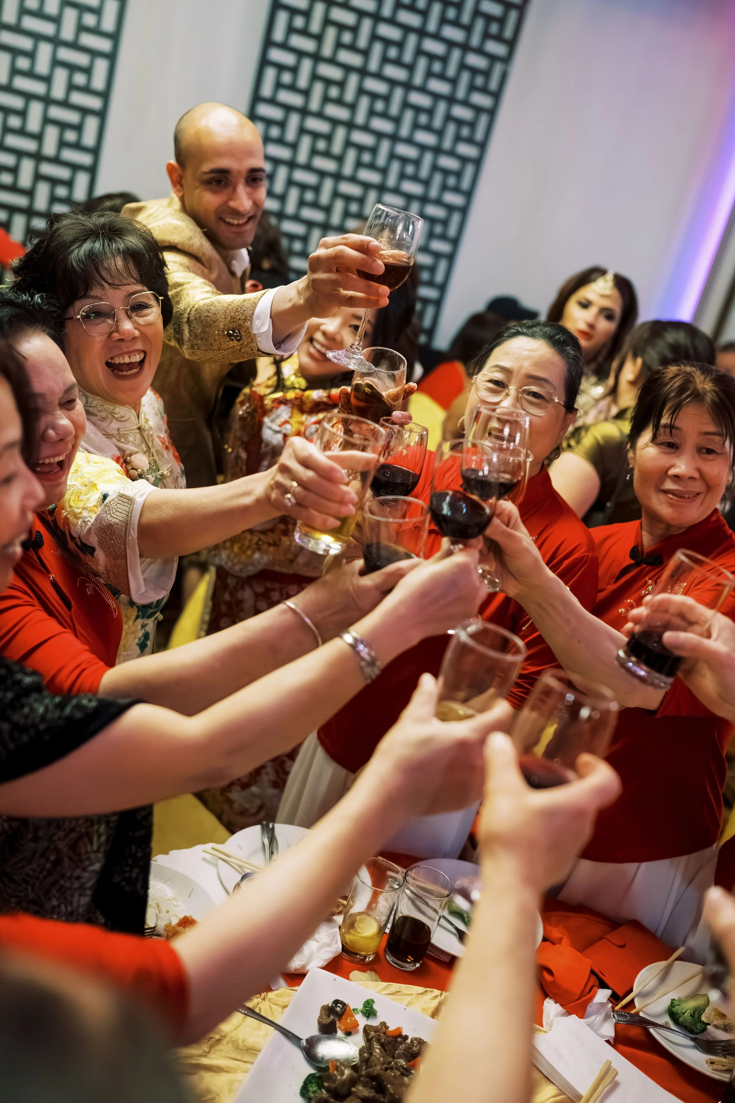 A group of people at a celebration or gathering, raising glasses of red wine for a toast, dressed in colorful and traditional attire, with food on the table and smiling faces.
