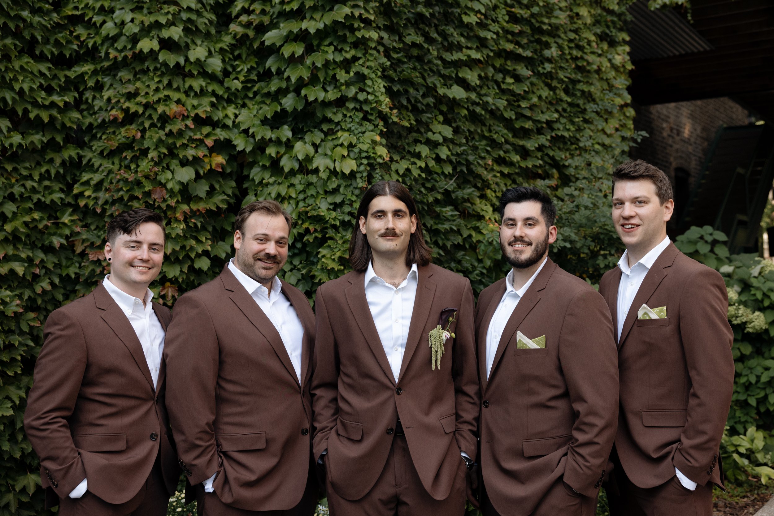 Group of five men in brown suits standing outdoors in front of a green ivy wall.
