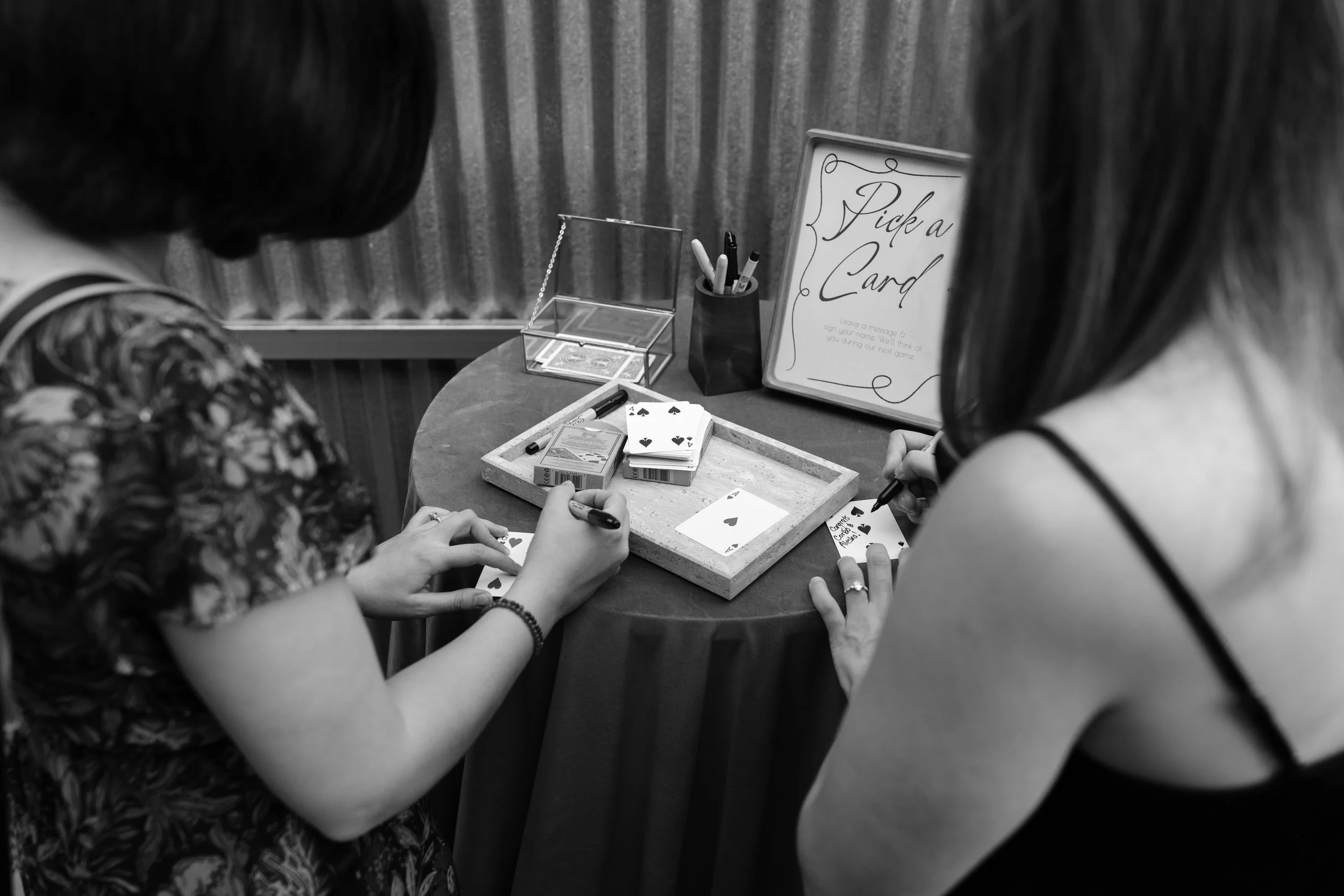Two women playing a game of cards at a table. One is holding an ace of spades, and the other appears to be writing on a card. A sign on the table instructs to pick a card and leave a message.