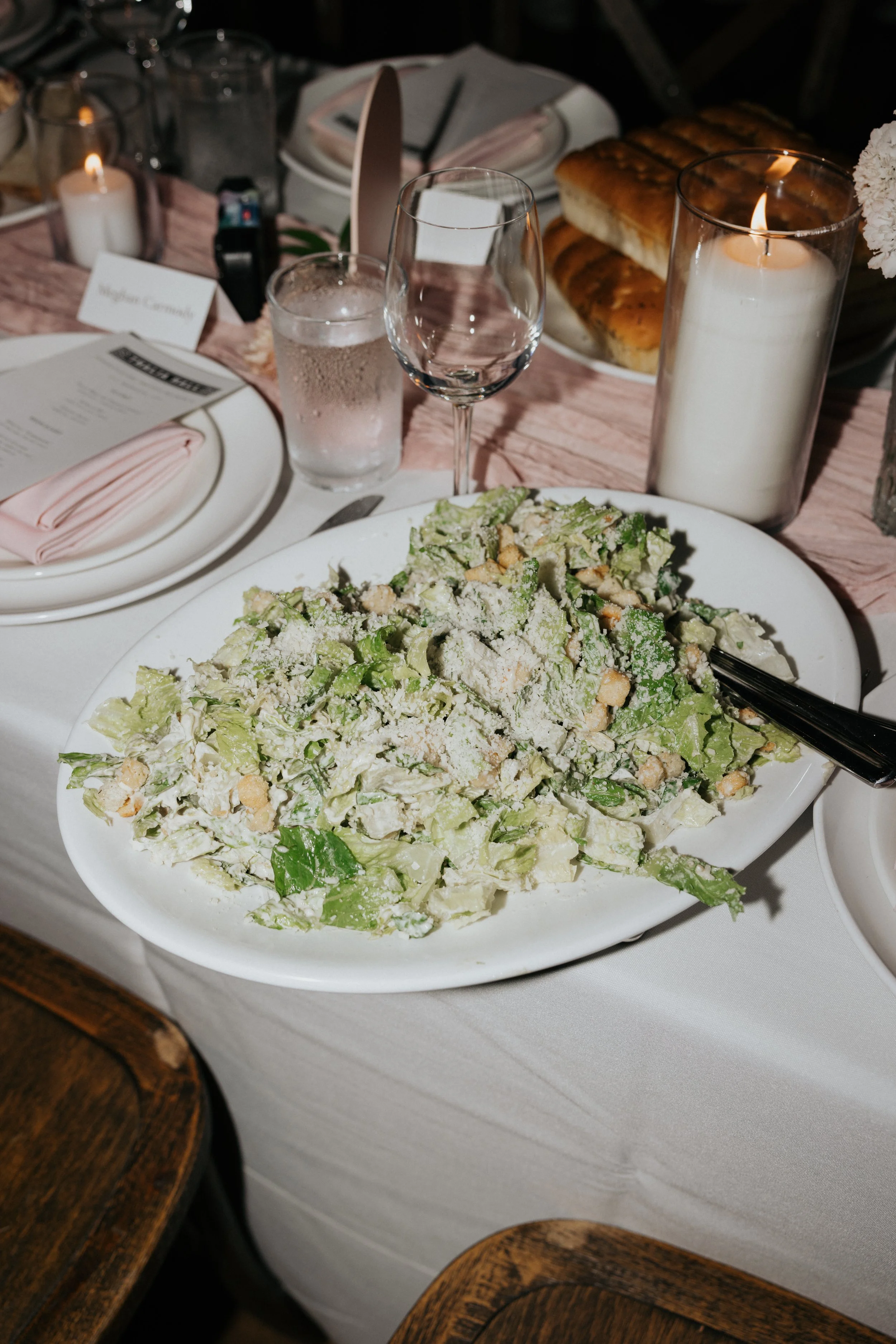 A dining table with a large Caesar salad on a white oval plate, bottles of candles, glasses, bread, and a menu, set with a pink tablecloth.