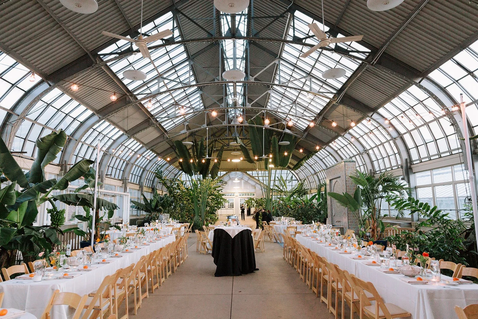 A large indoor event space with a glass ceiling and abundant greenery. Long banquet tables with white tablecloths and place settings are arranged along both sides. A few people are visible in the background near a decorative partition. String lights 