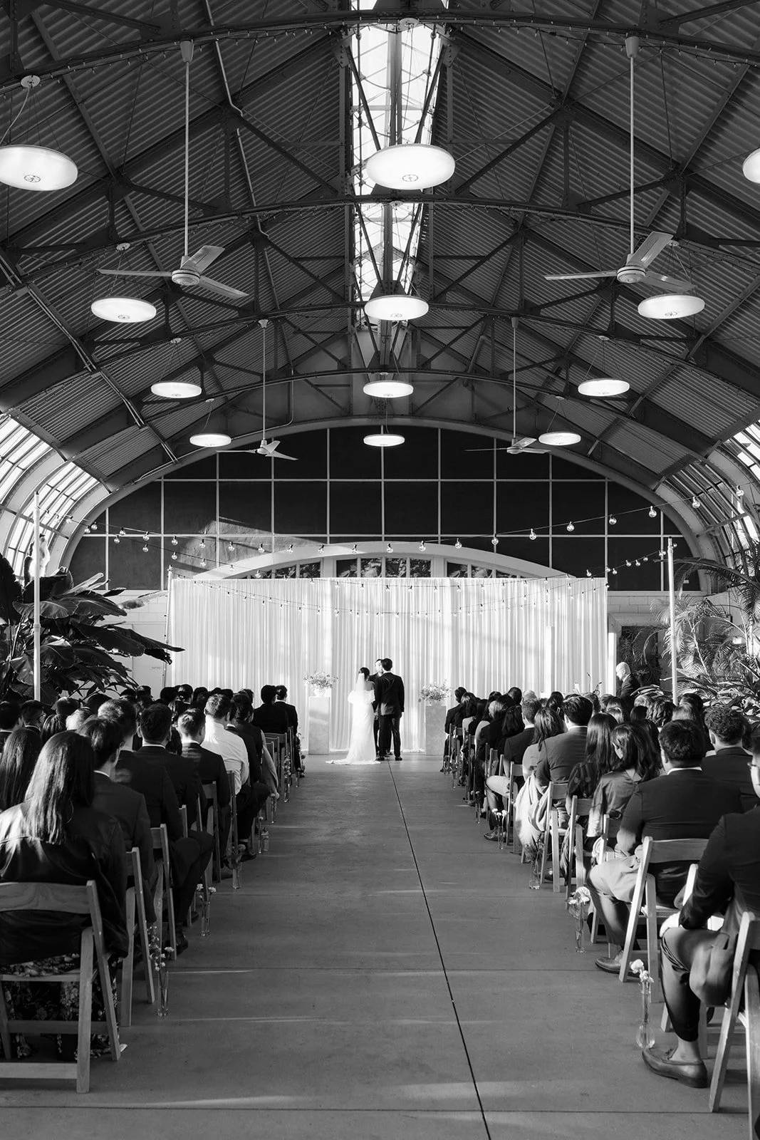 A wedding ceremony taking place in a large, industrial-style venue with a high, curved metal ceiling. The bride and groom are standing at the front in front of a curtain backdrop, facing each other. Guests are seated on either side, watching the cere