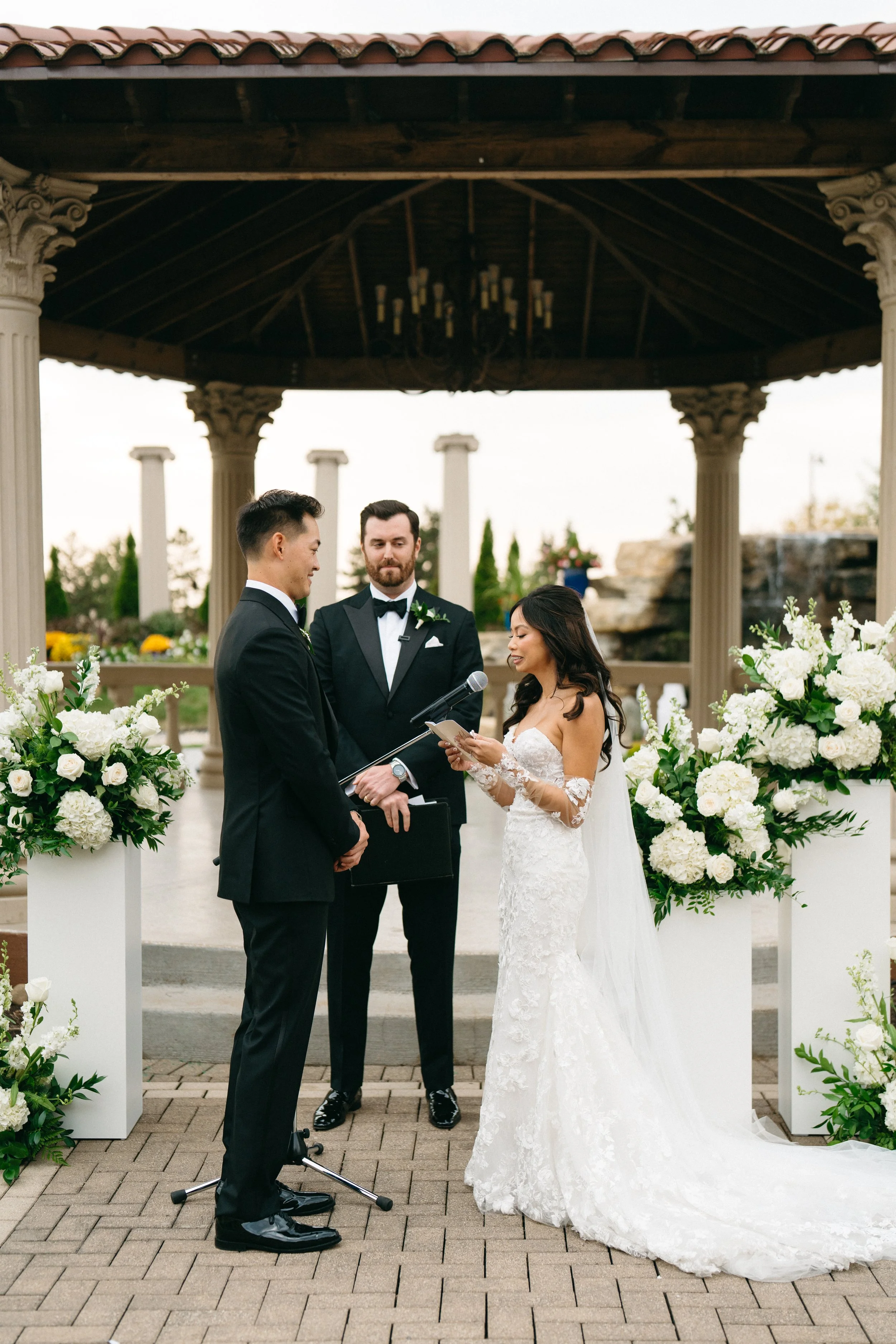 A bride and groom exchange vows at an outdoor wedding ceremony under a wooden pavilion with columns. The bride wears a white lace wedding dress with long sleeves, and the groom wears a black tuxedo. An officiant stands behind them holding a book, wit