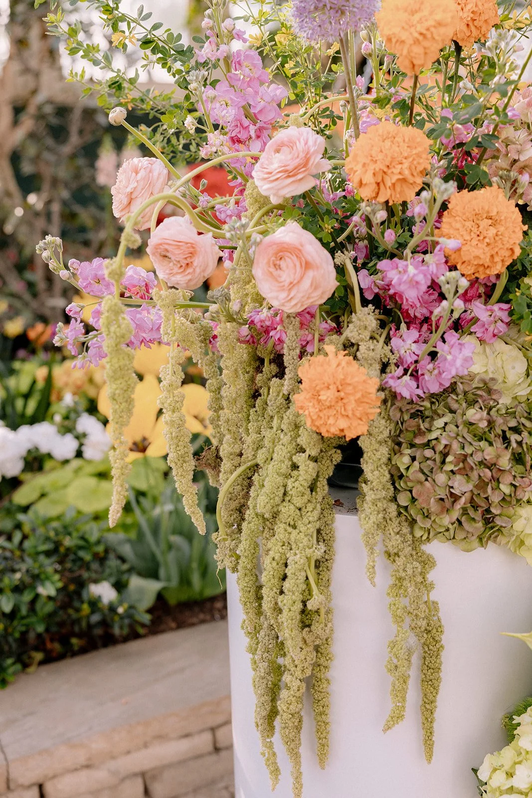 A floral arrangement in a white vase with pink ranunculus, orange marigolds, purple stock, and green hanging amaranthus.