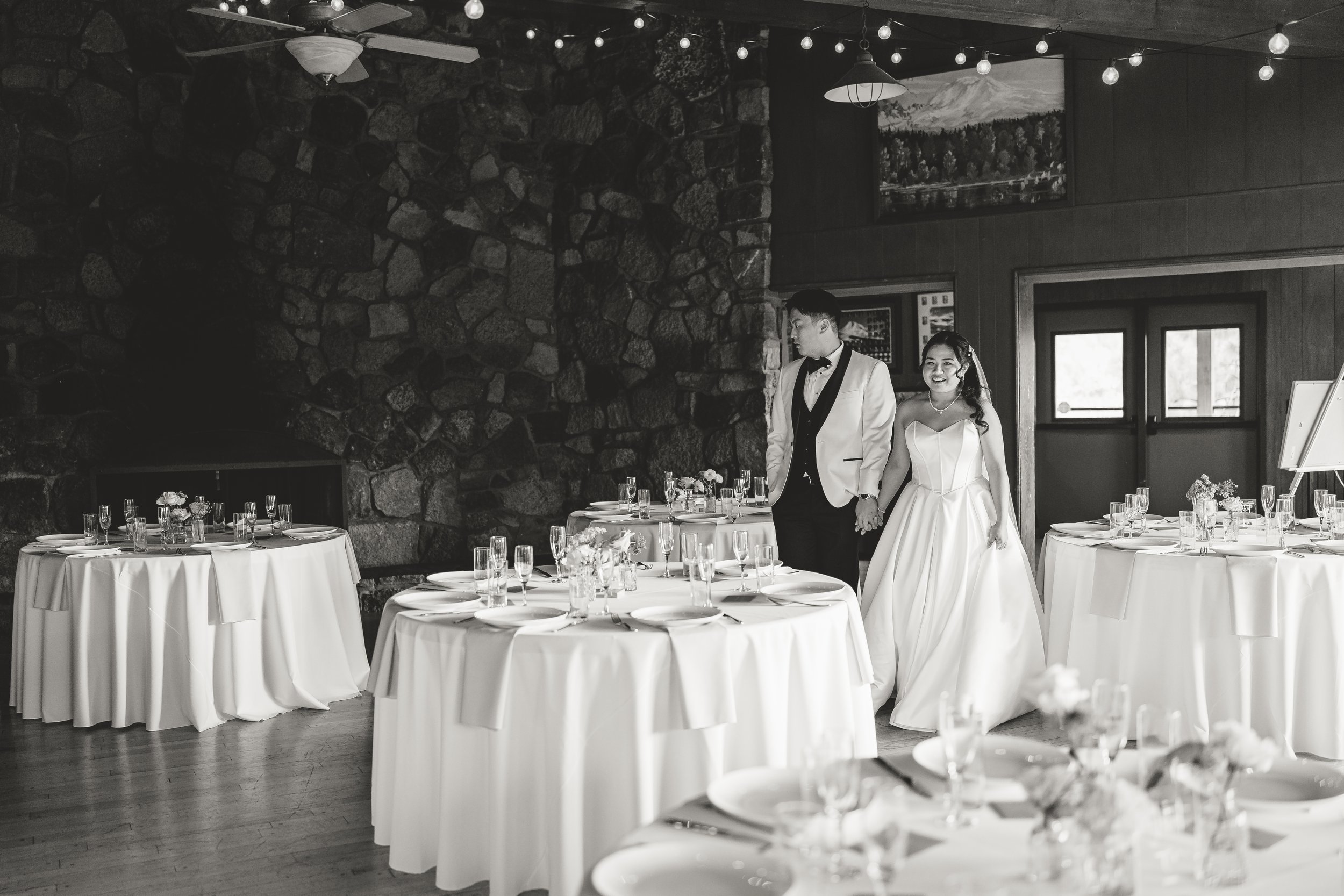A bride and groom holding hands inside a decorated reception hall with round tables, white tablecloths, glassware, and floral centerpieces, featuring a stone wall, window, and landscape painting in the background, with hanging string lights and a cei