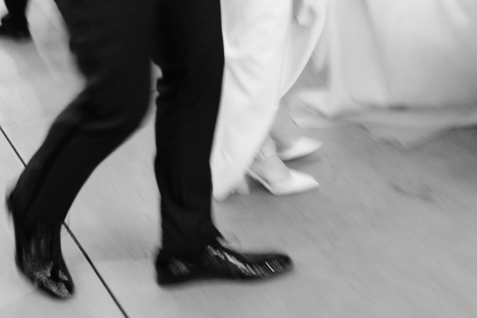 A person wearing shiny high-heeled shoes and dark pants is standing next to a white bed with a person in a white dress with their feet visible.