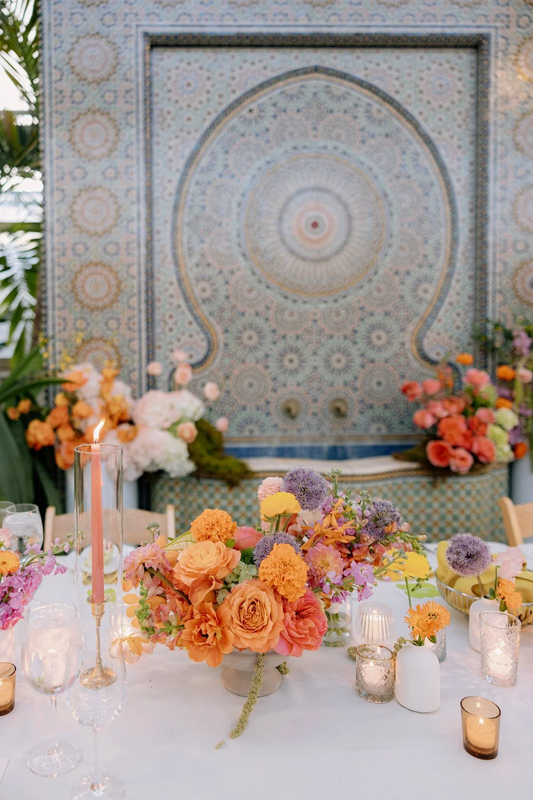 Elegant indoor event table setting with vibrant floral centerpieces, candles, and glassware, in front of a large, intricate mosaic wall.