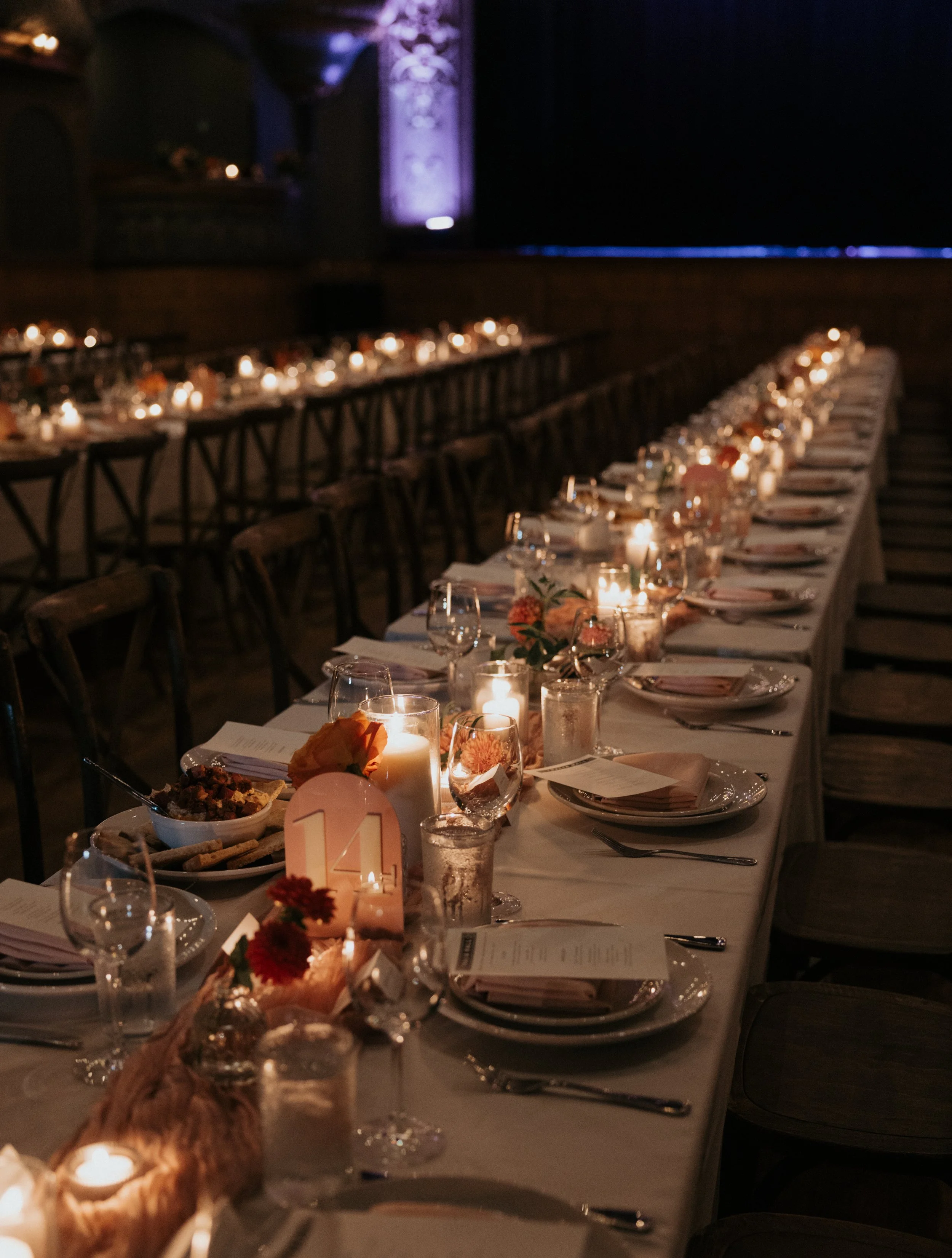 A long banquet table decorated with candles, flowers, glasses, and place settings in a dimly lit event space.