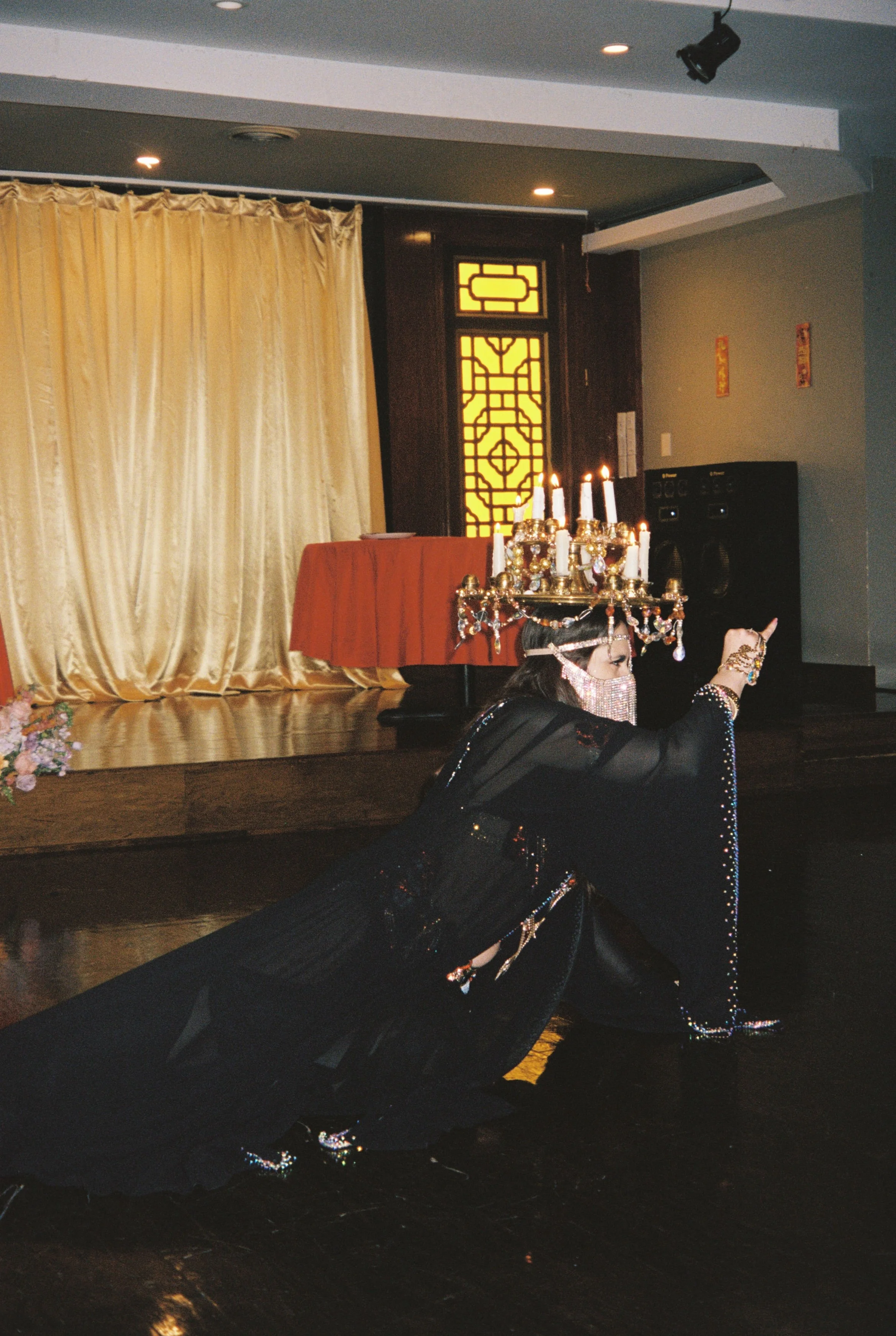 A woman dressed in traditional Asian attire with elaborate jewelry and a large decorative headpiece dances on a stage. The background features gold curtains, a table draped with a red cloth, and a decorative screen with yellow and black patterns. The