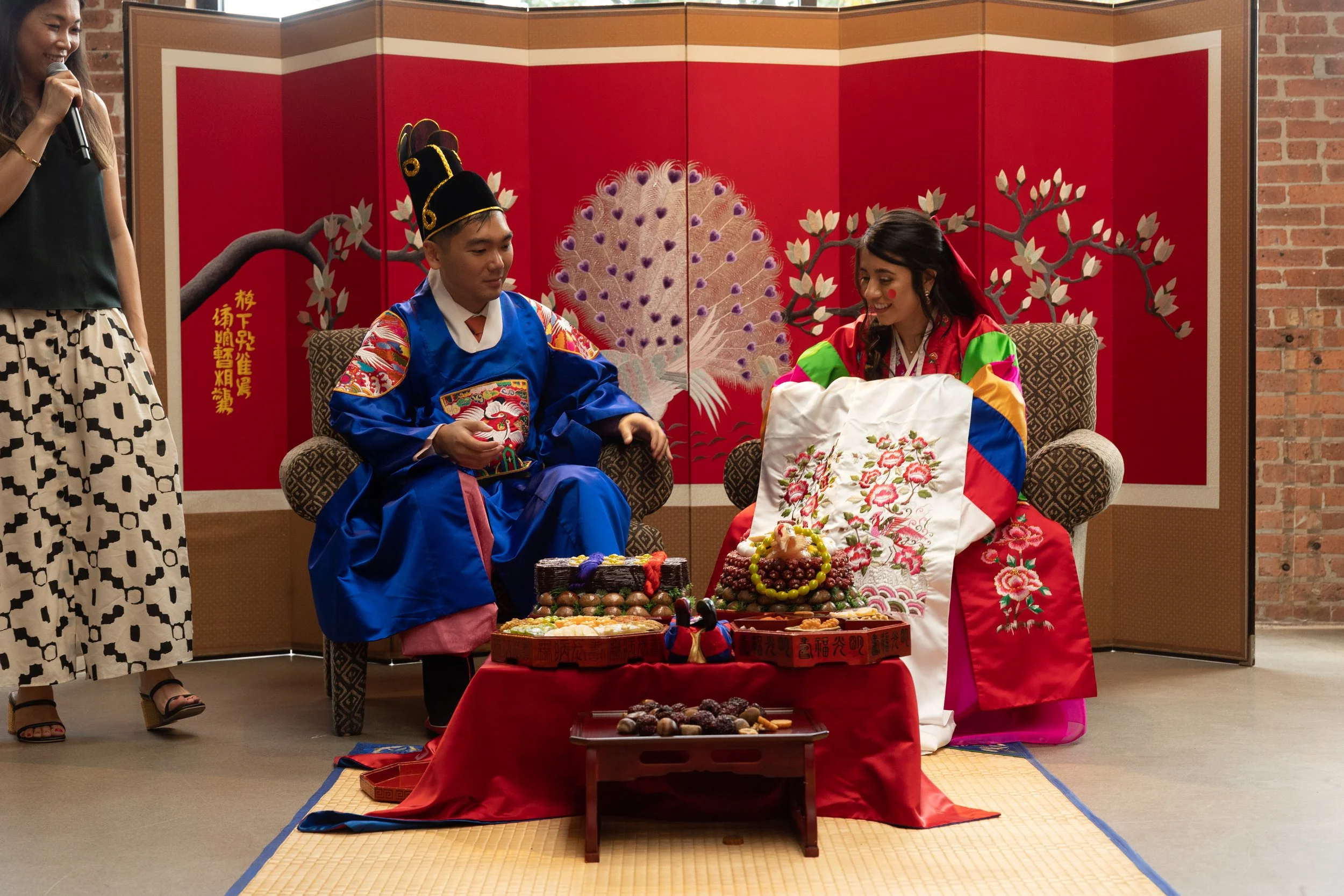 A traditional Asian ceremony with a man and a woman dressed in colorful traditional attire. The woman is holding a white embroidered cloth, and various ceremonial foods and offerings are displayed on a red table in front of them. A decorative red and