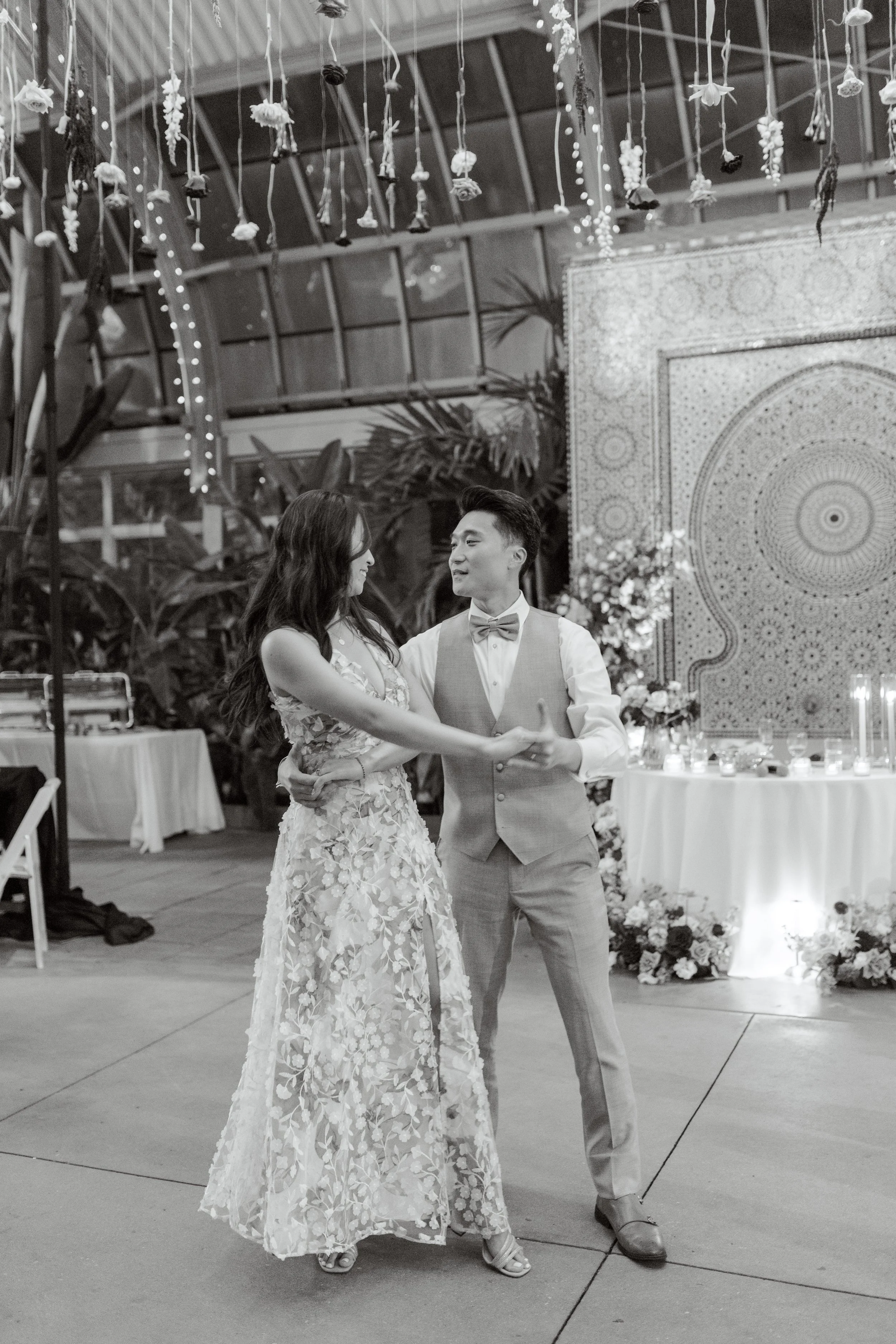 A couple dancing at a wedding reception, with decorations and a large patterned wall in the background.