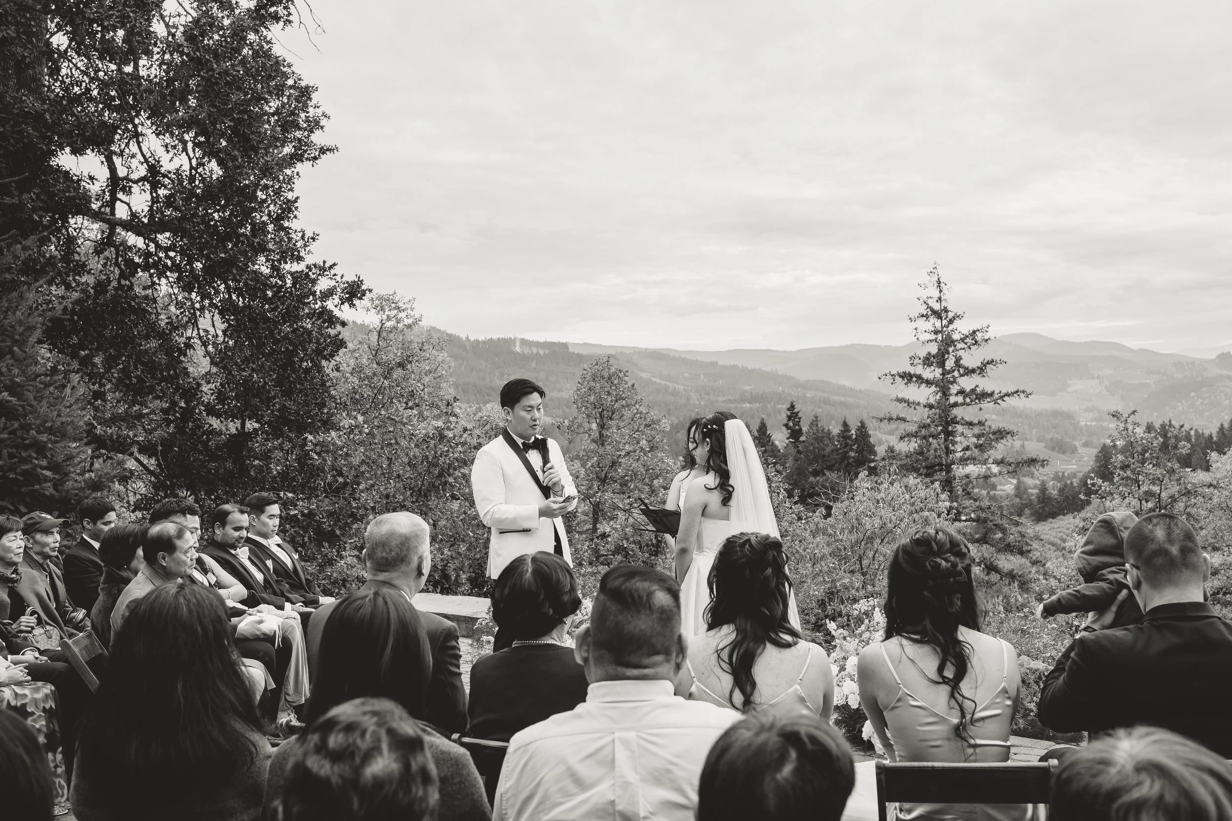 A black and white photo of a wedding ceremony outdoors with guests seated, a groom reading vows, and a bride listening, surrounded by trees and a scenic landscape in the background.