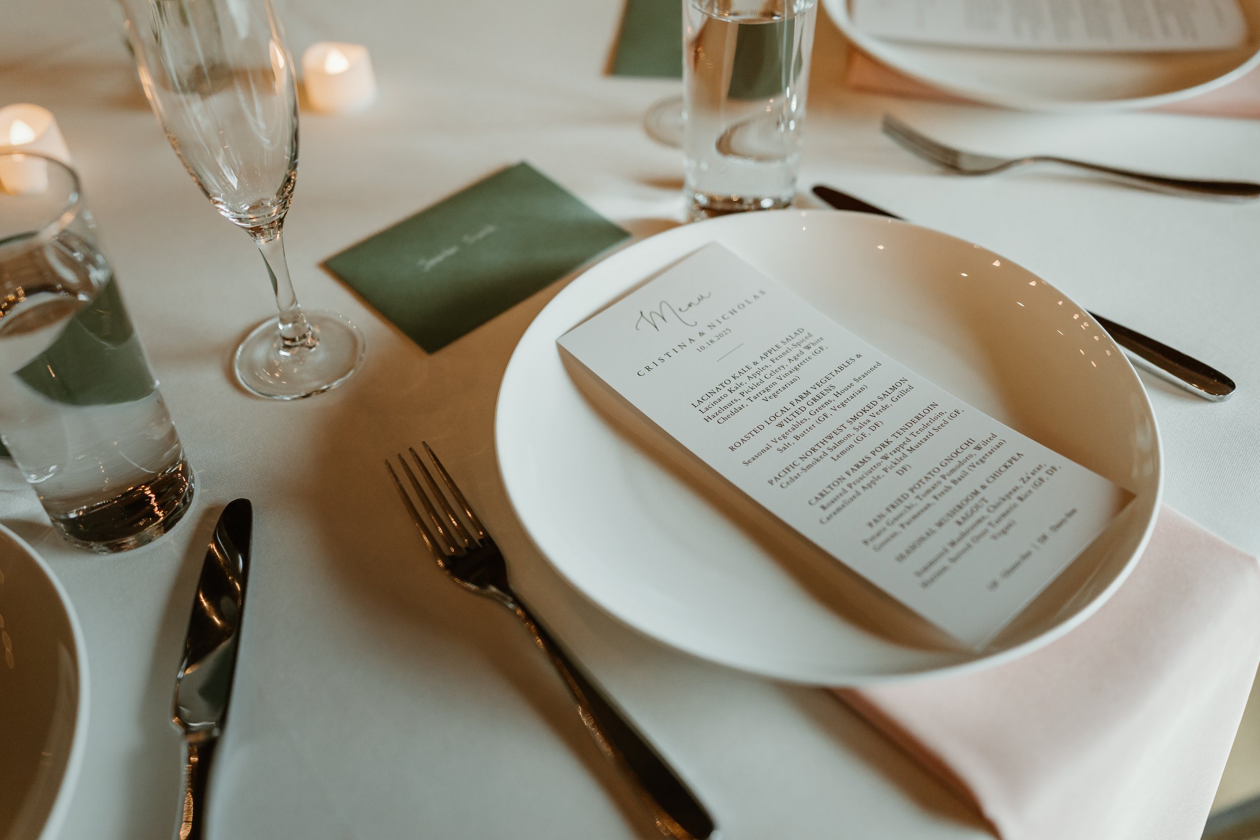 A table setting at a formal event including champagne glasses, a water glass, a fork, a knife, a spoon, a menu card, and a green place card. The menu has details of a meal, and the table is decorated with small candles.