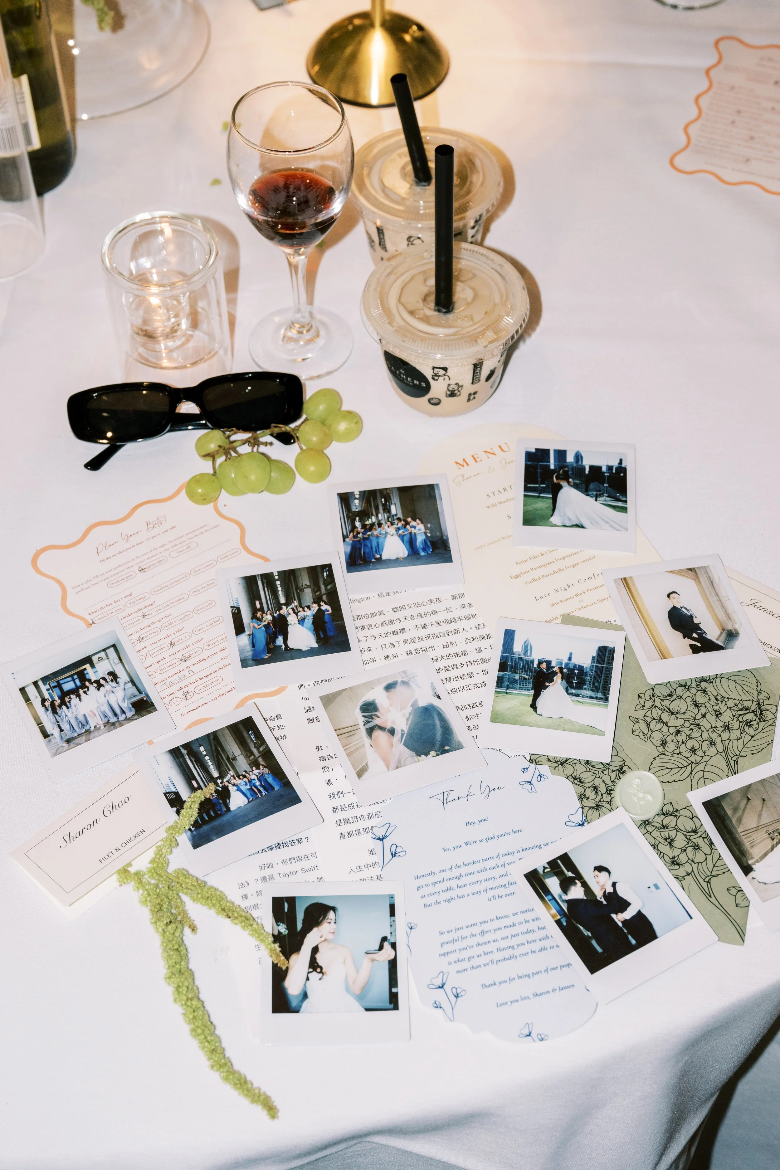 A table with wedding memorabilia including photographs, letters, a glass of red wine, iced coffee drinks with straws, sunglasses, a bunch of green grapes, a small candle, and some documents.