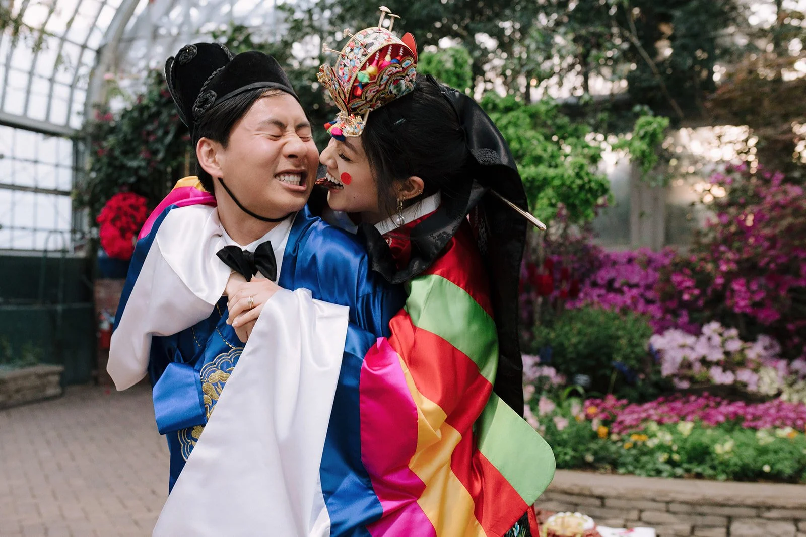Two people in traditional and colorful costumes sharing a joyful hug outdoors, surrounded by flowers and greenery.