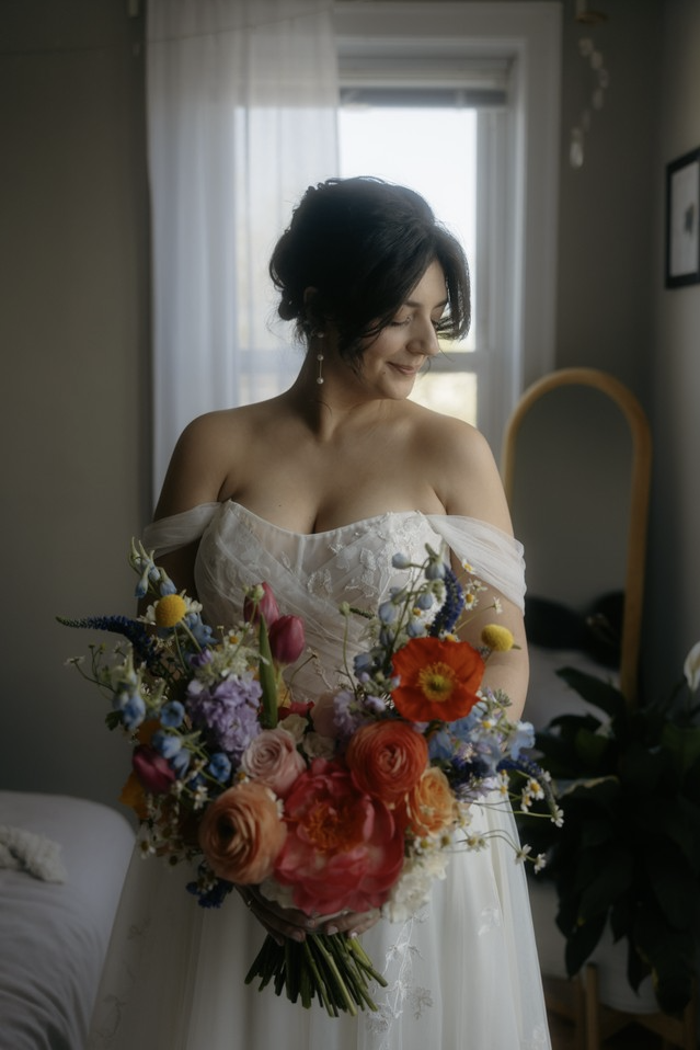 A bride with short dark hair in a white wedding dress holding a colorful bouquet of flowers inside a room with a window in the background.