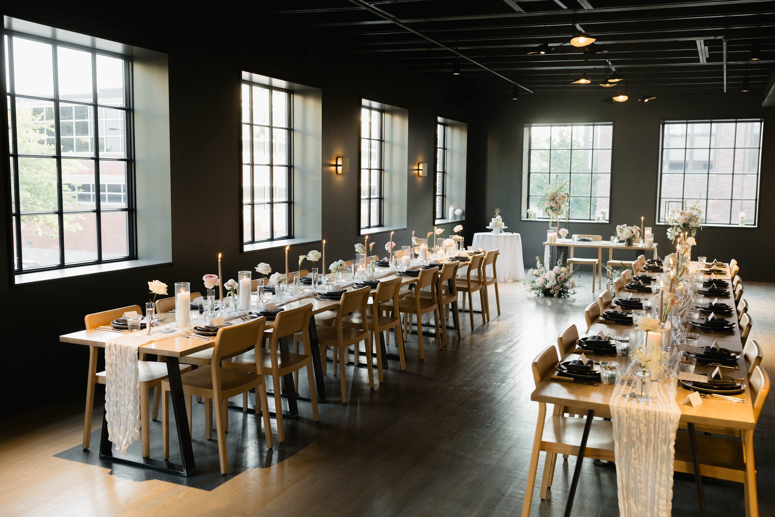 Elegant event space with long dining tables decorated with white flowers, candles, and black dinnerware, set for a formal gathering in a room with large windows and black ceiling panels.