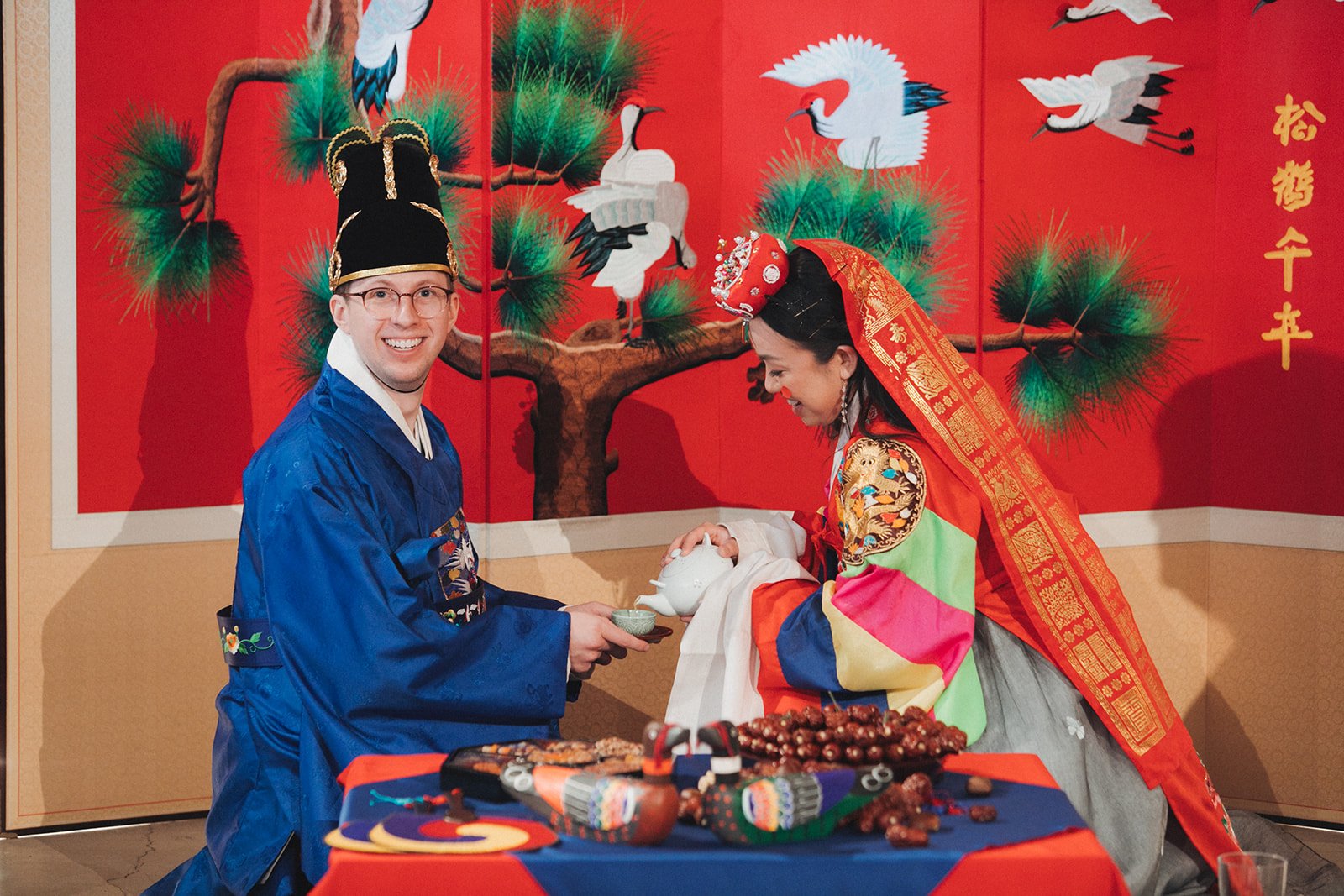 People in traditional Chinese clothing exchanging tea in front of a red background with cranes and pine trees, indicating a Chinese cultural ritual.