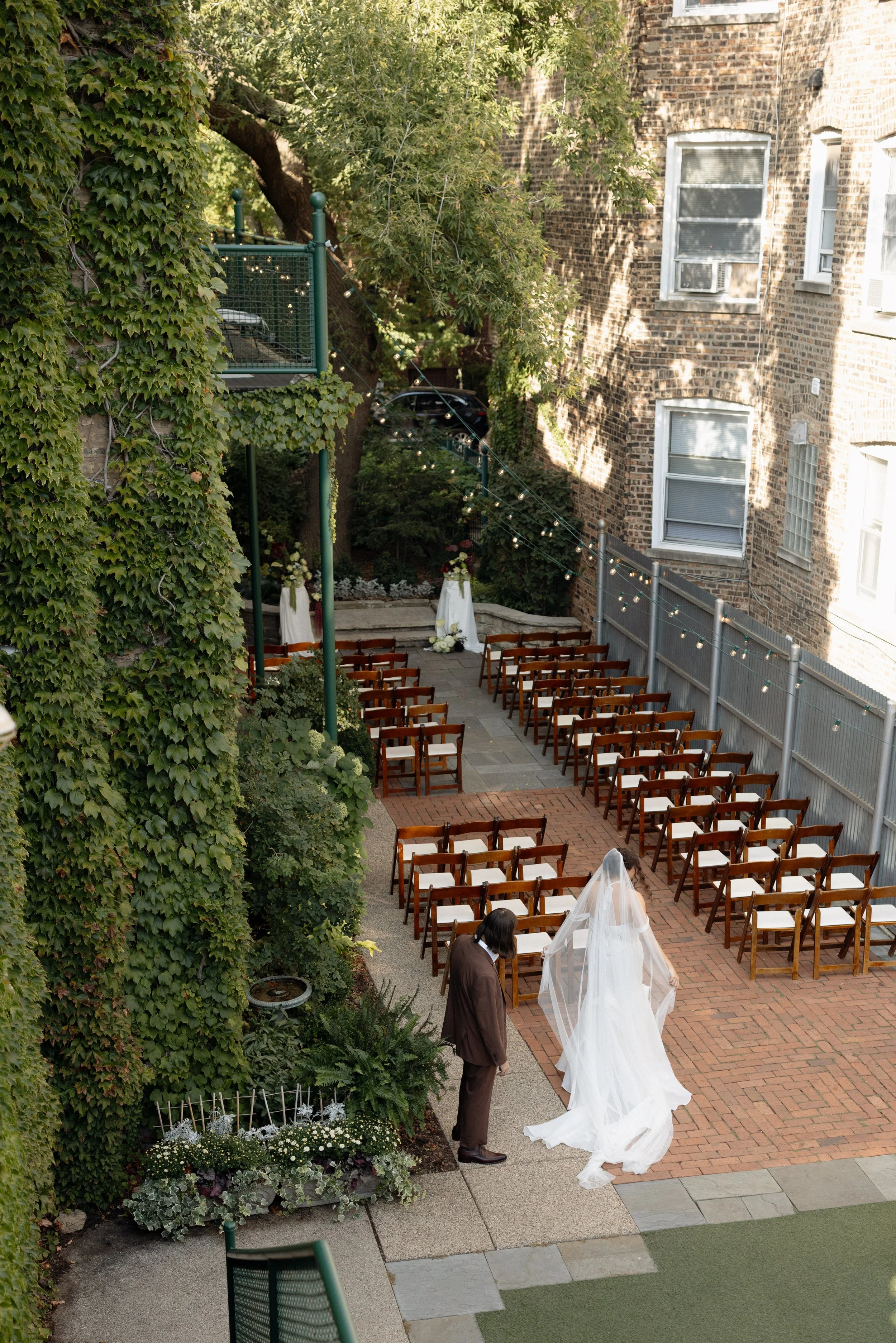A wedding ceremony setup in an outdoor courtyard with wooden chairs, a white cloth altar, and floral arrangements. A person in a brown suit is standing near a bride in a white wedding dress, who is adjusting her veil. The courtyard has a brick pathwa