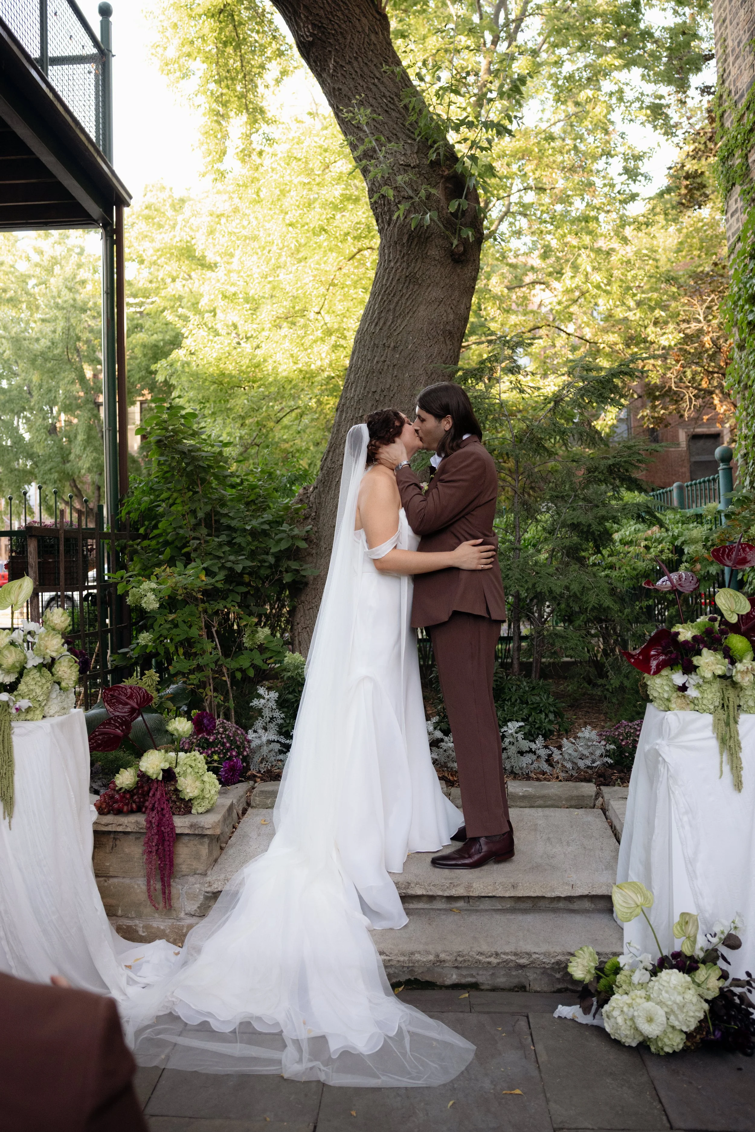 A bride and groom sharing a kiss during their outdoor wedding ceremony, surrounded by greenery and flowers. The bride is in a white wedding gown with a veil, and the groom is in a brown suit.