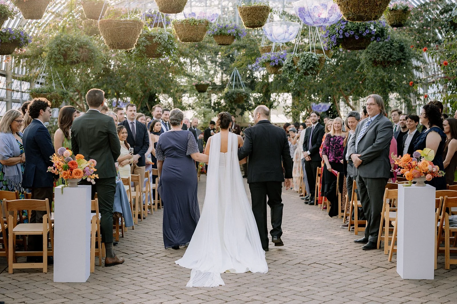 A wedding ceremony taking place outdoors inside a greenhouse. The bride, dressed in a white wedding gown, is walking with two attendants down the aisle, while guests are standing and watching on both sides. Hanging flower arrangements and floral disp