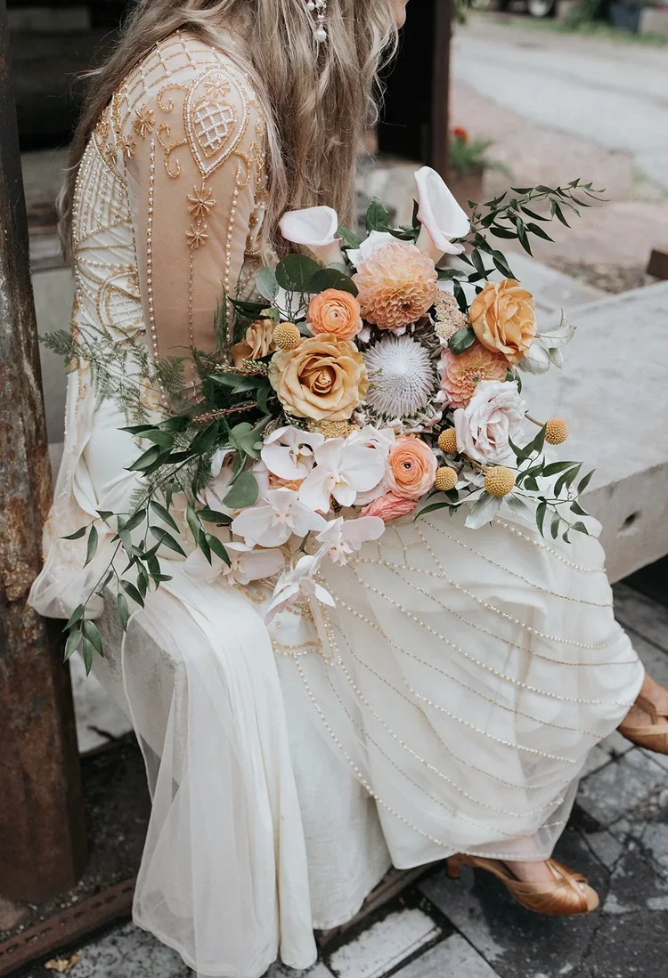 A woman sitting outdoors holding a large bouquet of peach, white, and cream-colored flowers with greenery, wearing a white dress with intricate gold embroidery on the sleeves.
