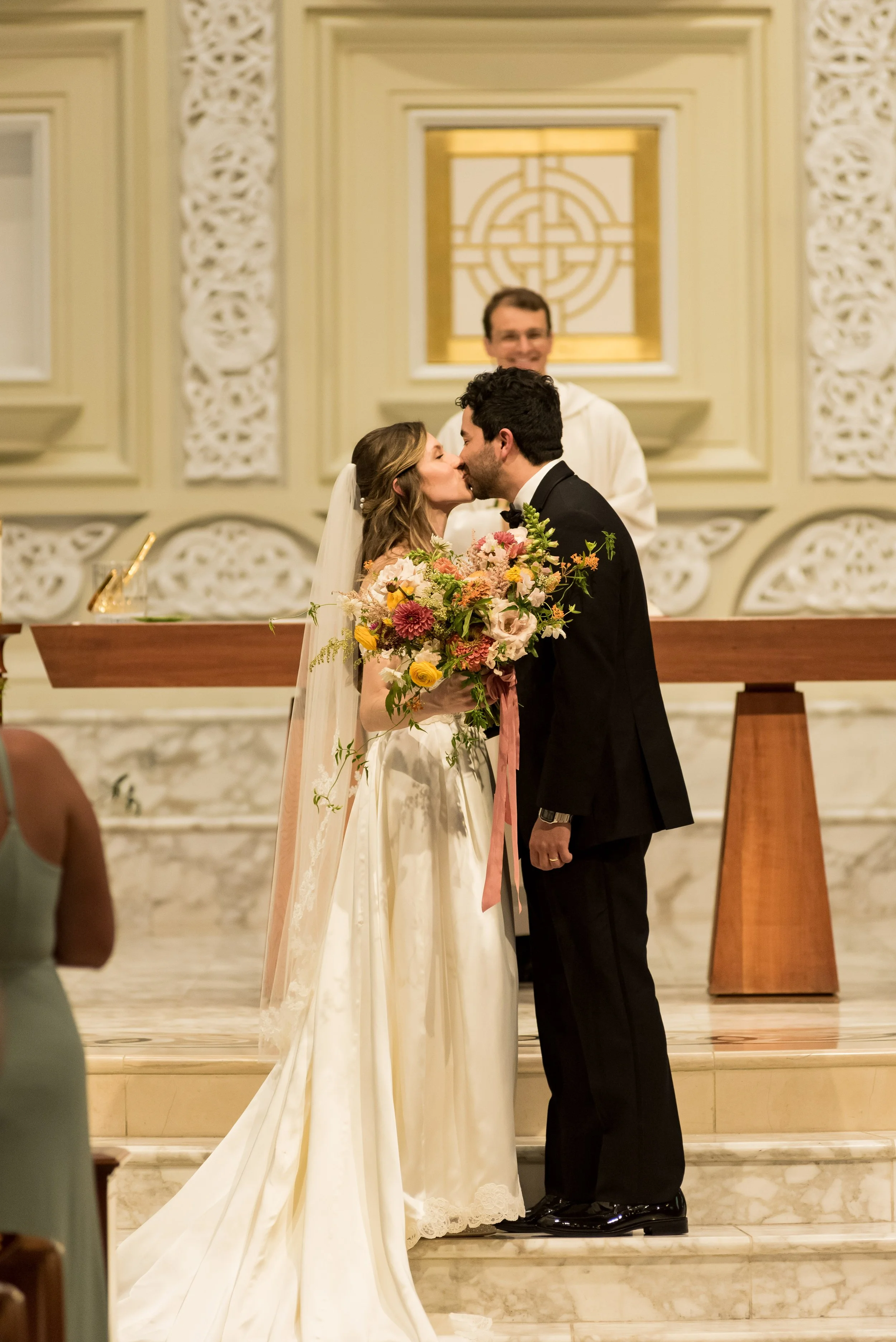 A bride and groom kiss at their wedding ceremony inside a church, standing on steps with a large bouquet of flowers. A man in white robes officiates behind them, in front of decorated walls with ornate designs.