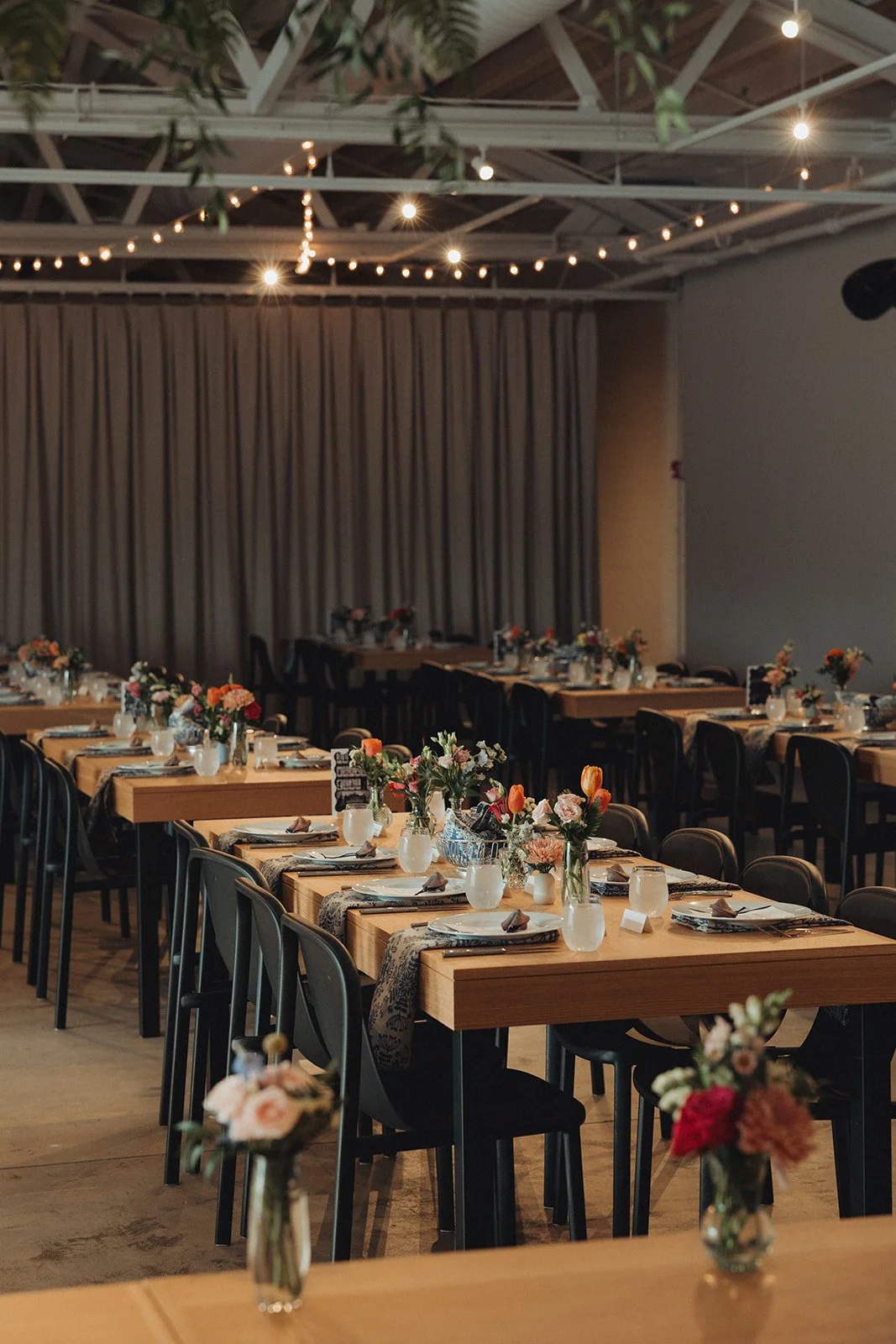 Indoor event space with long wooden tables set for a formal meal, decorated with vases of colorful flowers, napkins, and tableware. String lights hang from the ceiling, creating a warm, festive atmosphere, with a closed curtain in the background.