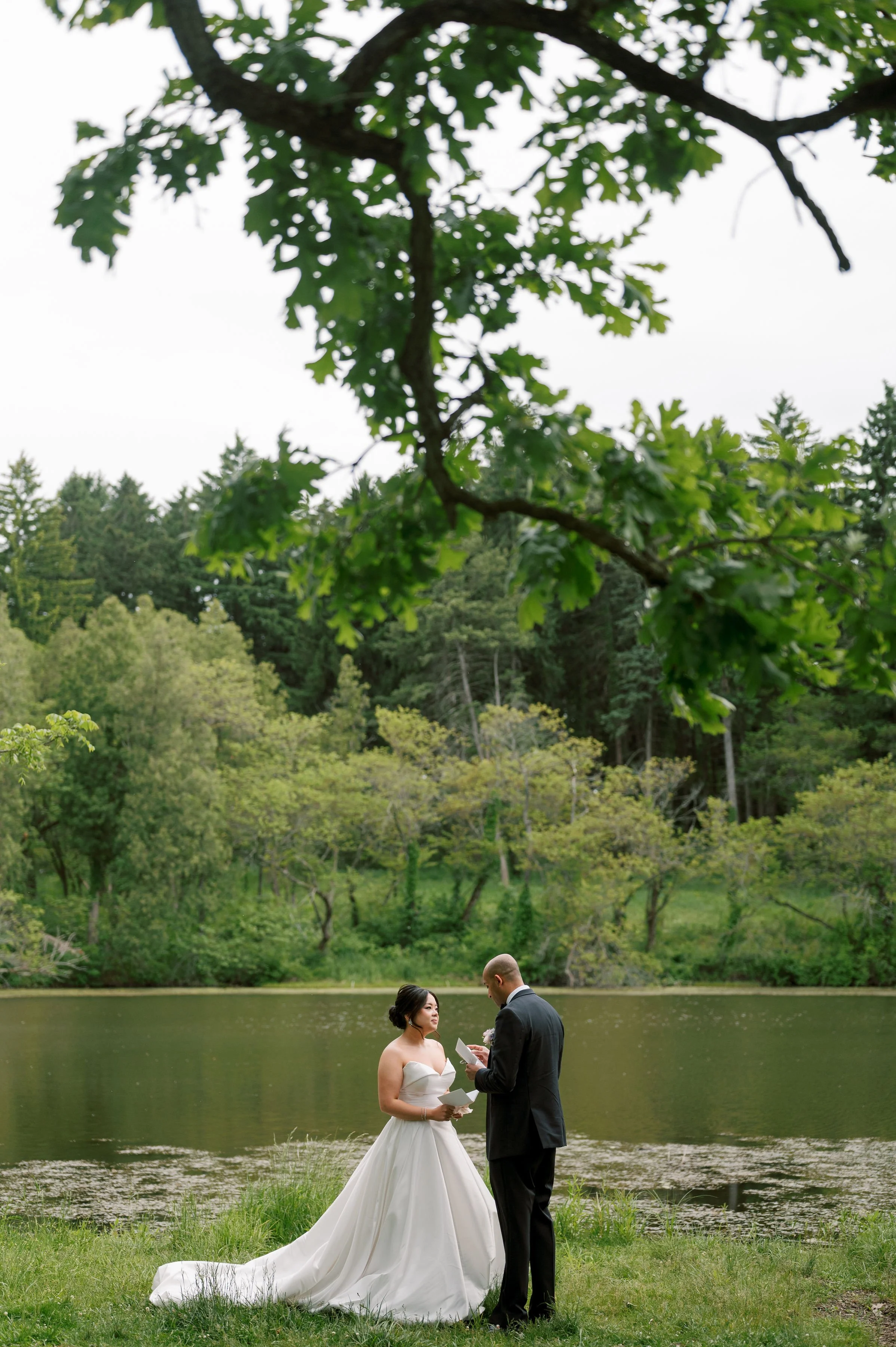 A bride and groom exchanging vows beside a lake surrounded by trees with an overcast sky.