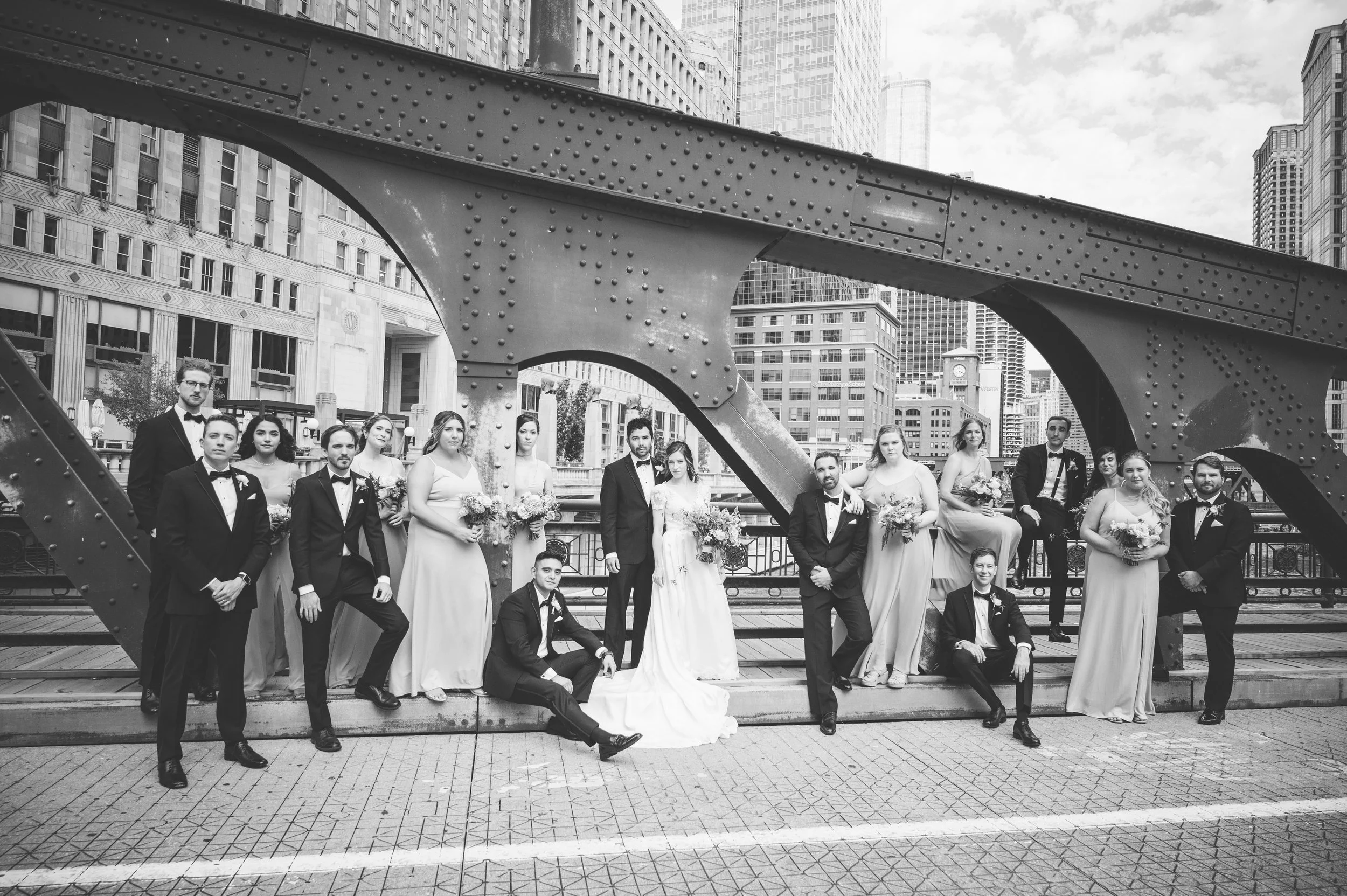 Black and white photo of a wedding party on a bridge in an urban city, with tall buildings in the background. The group includes men in tuxedos and women in white dresses, holding bouquets. They are posing for a group photo under a large, riveted ste