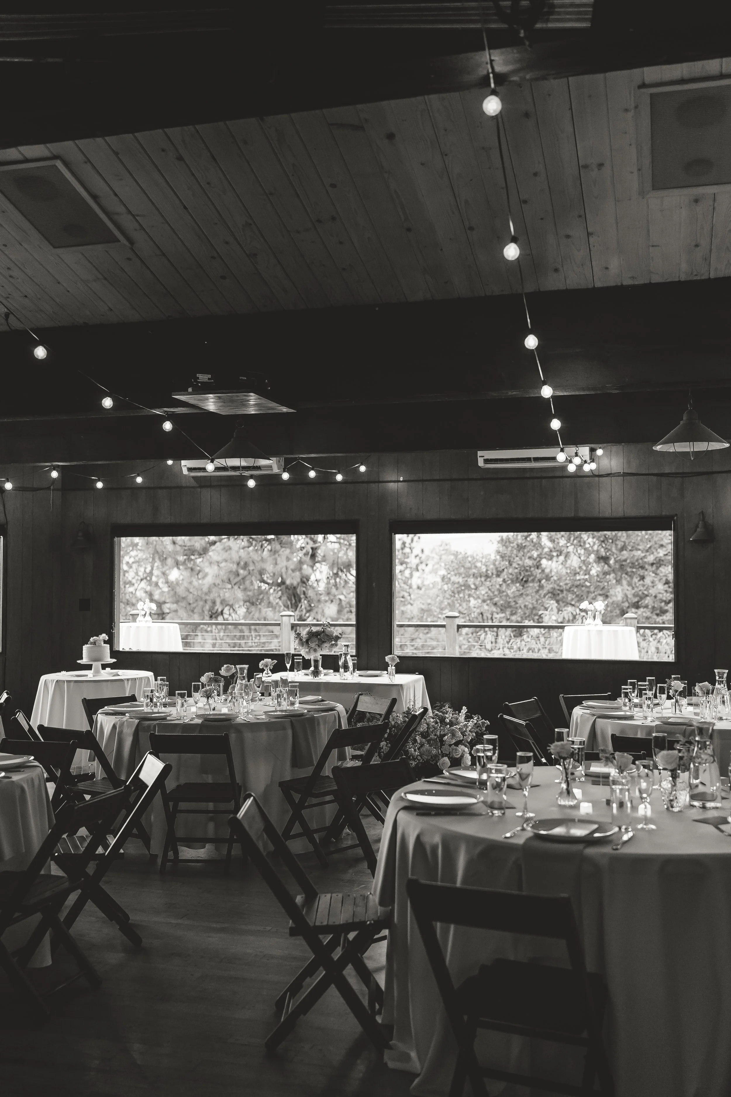 Interior of a decorated event space with round tables covered in white tablecloths, set with glasses, plates, and centerpieces, chairs around the tables, large windows showing outdoor scenery, and string lights hanging from the ceiling.