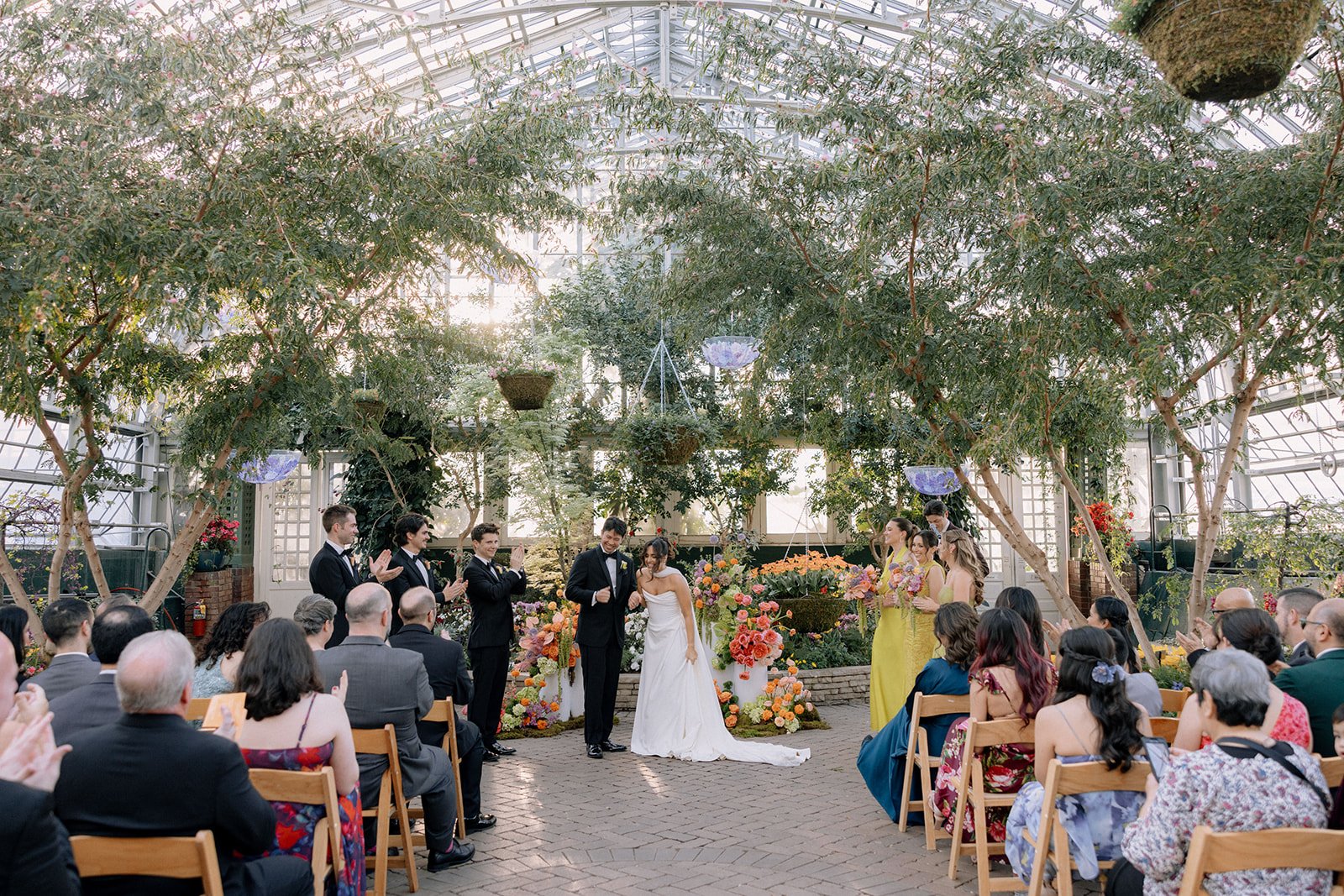 A wedding ceremony taking place inside a greenhouse with a glass ceiling. The bride and groom are at the center, surrounded by a bridal party and guests. The ceremony is decorated with vibrant flowers and lush greenery.