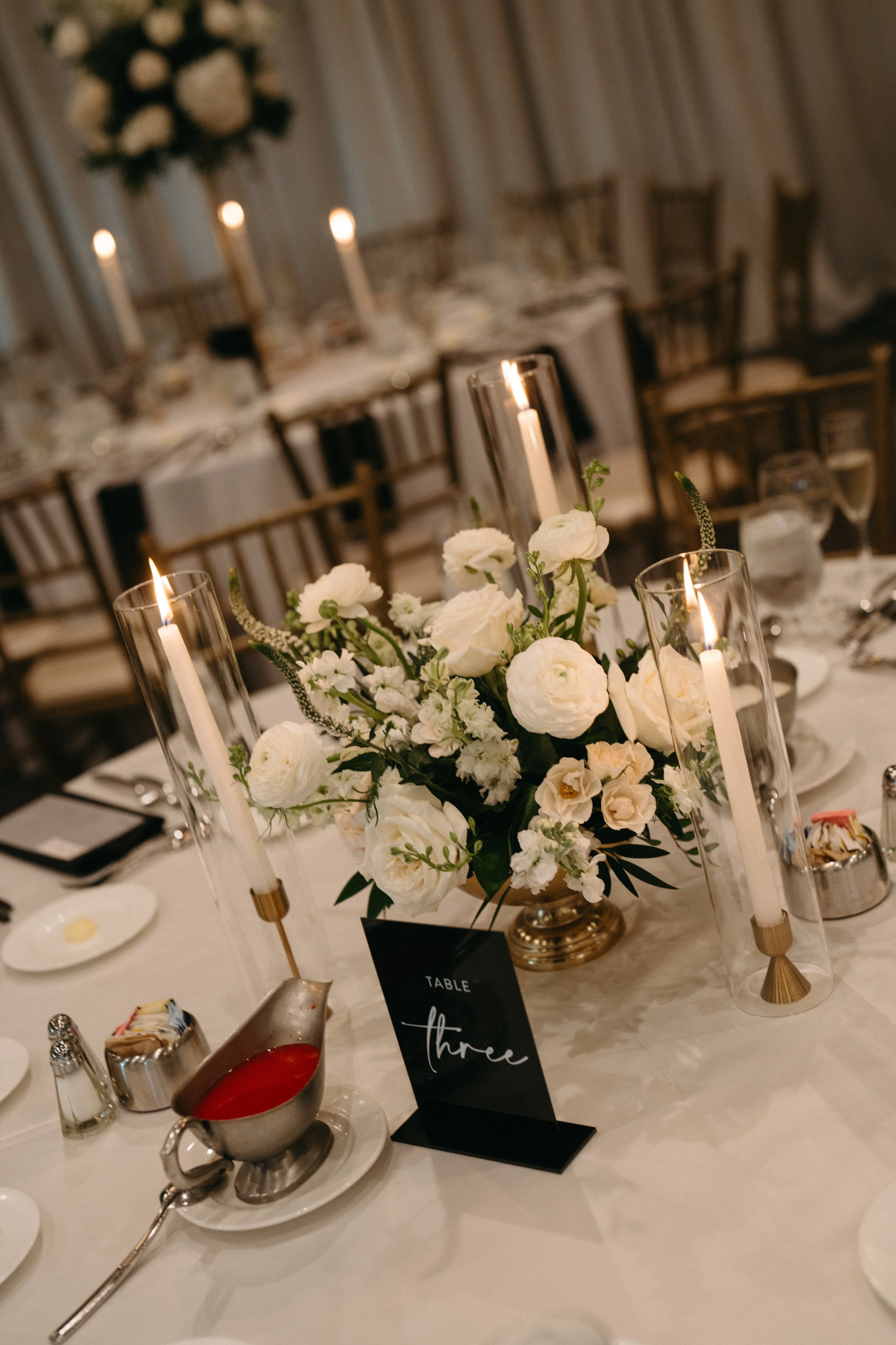 Elegant wedding reception table with white floral centerpiece and tall candles, set in a decorated venue with chairs and a backdrop in the background.