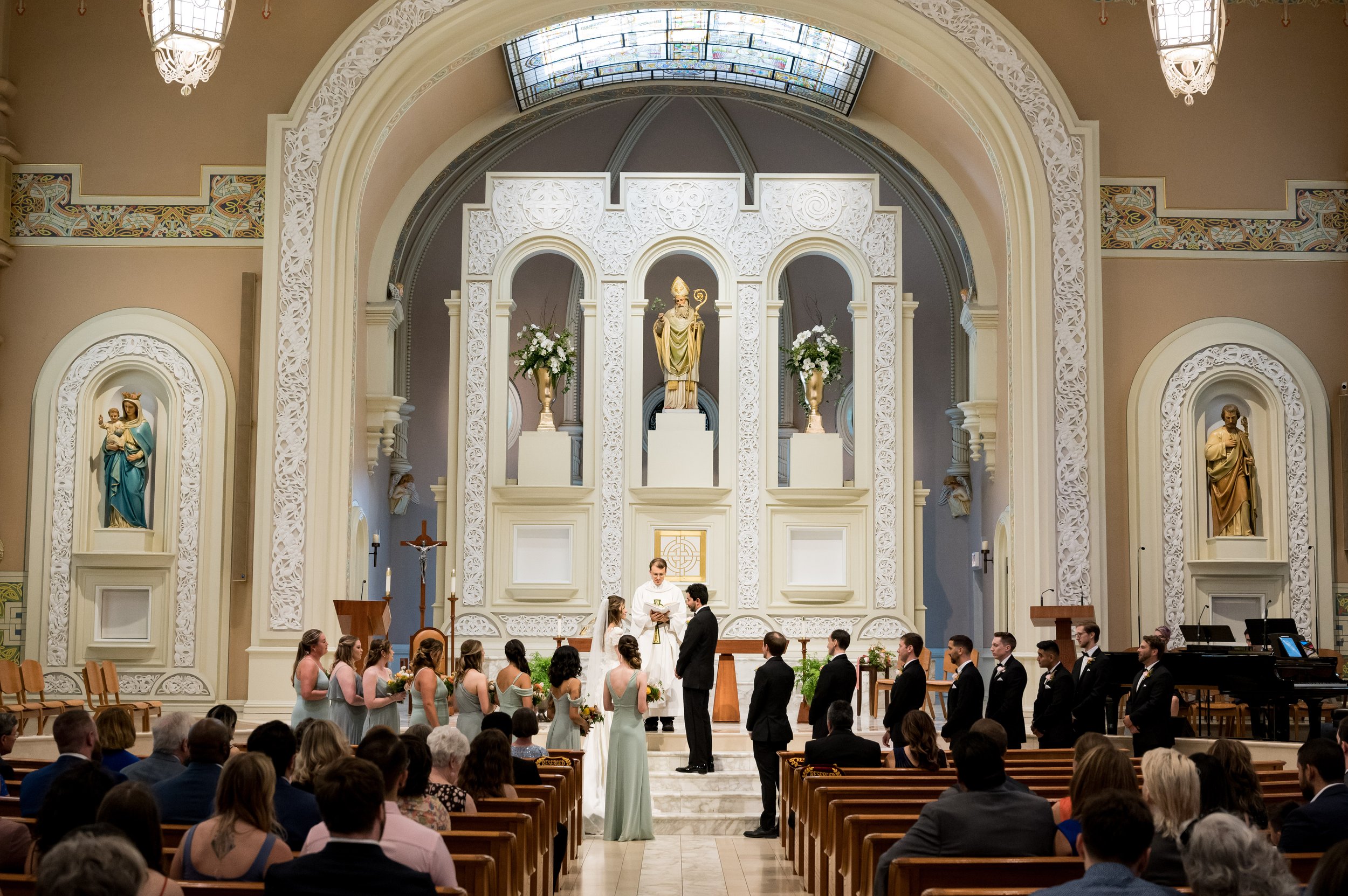 A wedding ceremony inside a church with a group of bridesmaids and groomsmen standing before the altar. The altar has religious statues and stained glass windows. The congregation is seated watching the event.