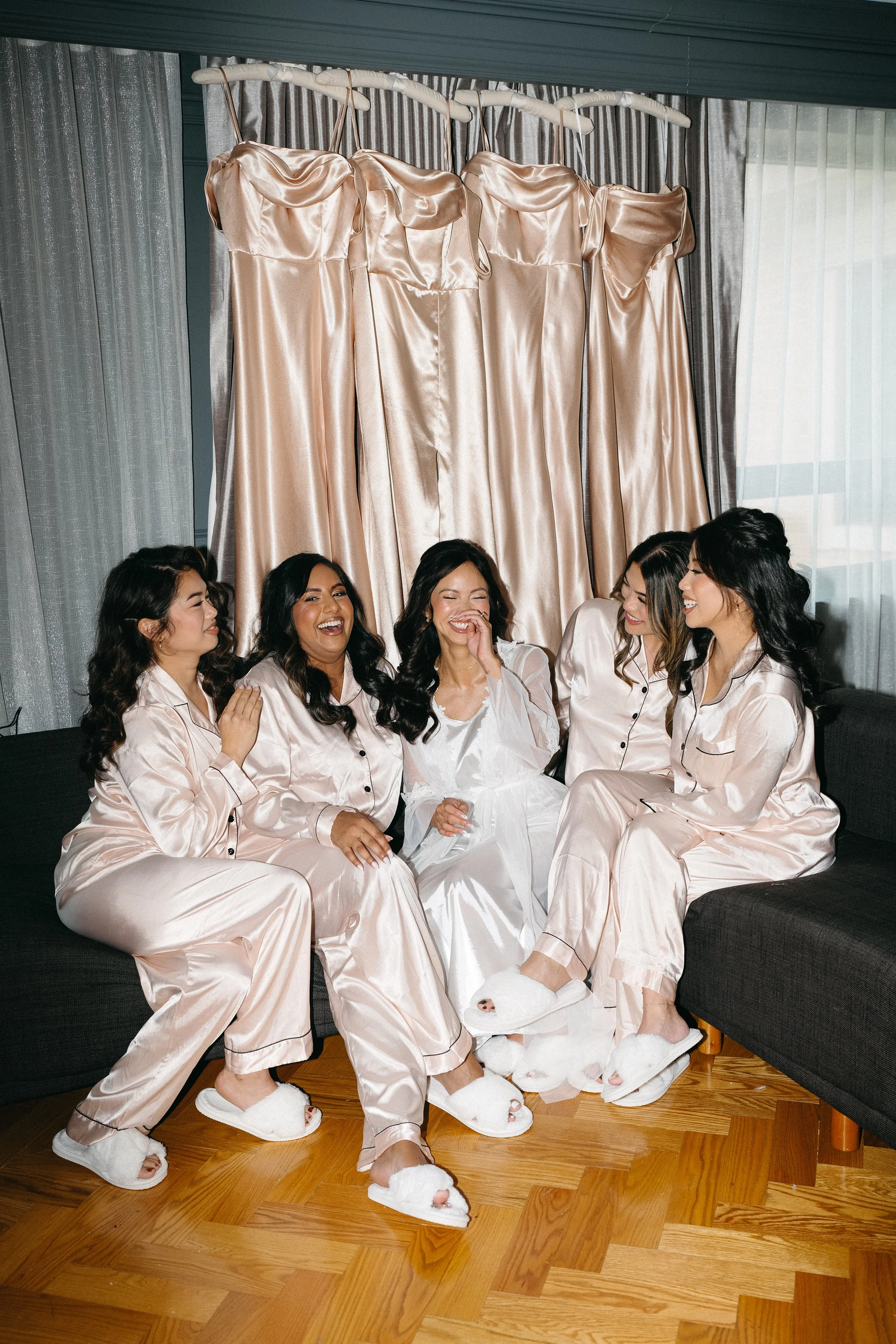 Group of women in satin pajamas and fluffy slippers sitting on a couch, laughing and enjoying each other's company in a living room with a window and beige curtains in the background.