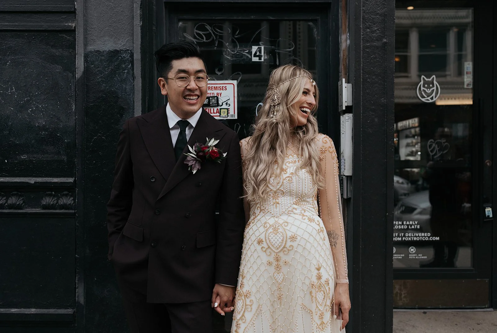 A happy couple, a man in a dark suit with glasses and a woman in a gold and cream embroidered gown, holding hands and smiling, standing outside a black storefront.