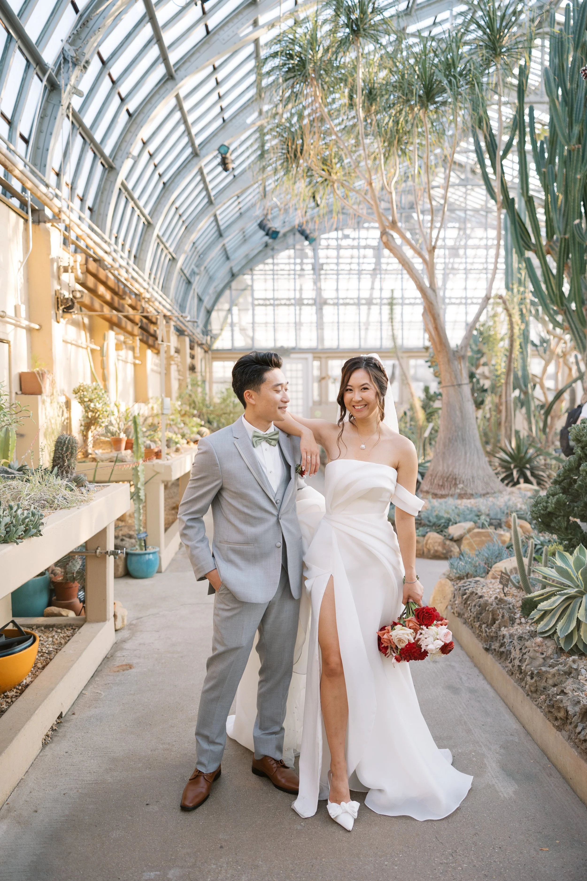 A bride and groom walking inside a greenhouse, with tropical plants and cacti around them. The bride is wearing a white strapless wedding gown and carrying a bouquet, while the groom is in a light gray suit with a green bow tie.