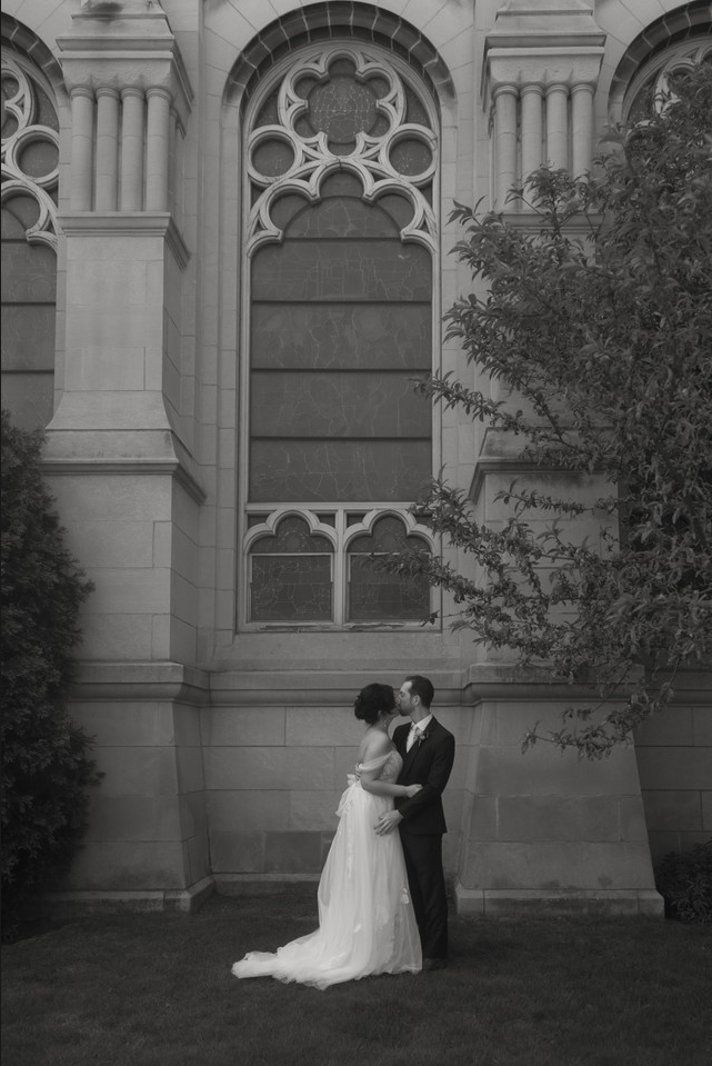 A bride and groom stand close together in front of a historic stone building with large stained glass windows. The bride wears a long white wedding dress, and the groom is dressed in a tuxedo. They are kissing or about to kiss, surrounded by a grassy