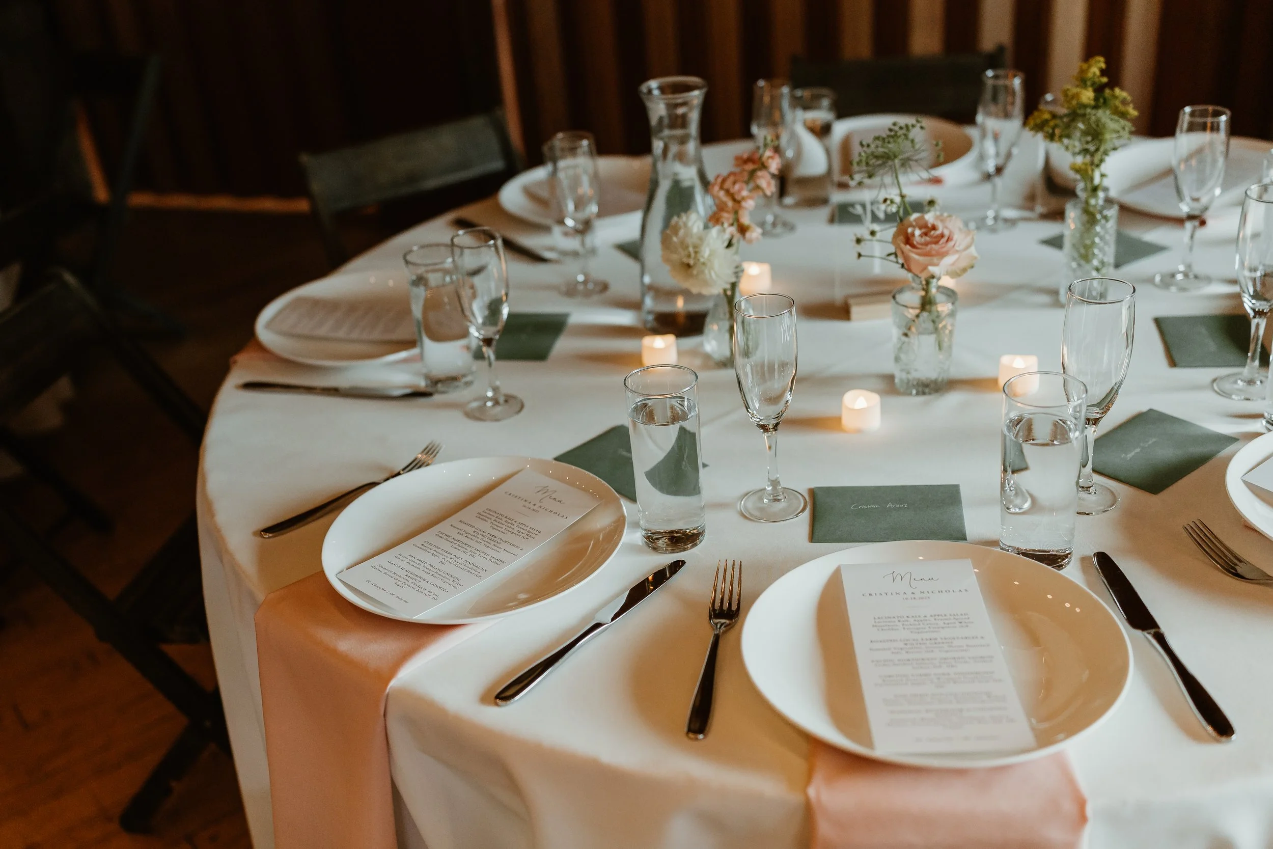 Table set for a formal event with white tablecloths, floral centerpieces, candles, plates, glasses, and silverware.