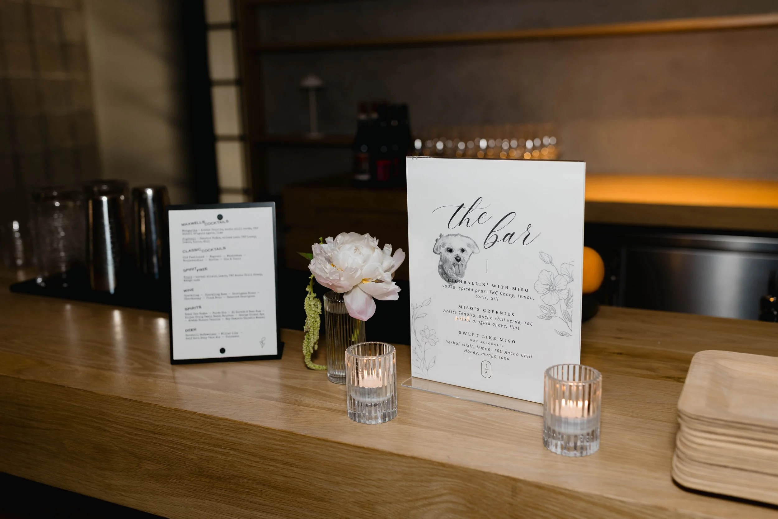 A bar counter with a floral arrangement, cocktail menus, and candles, with a dark background and shelves.