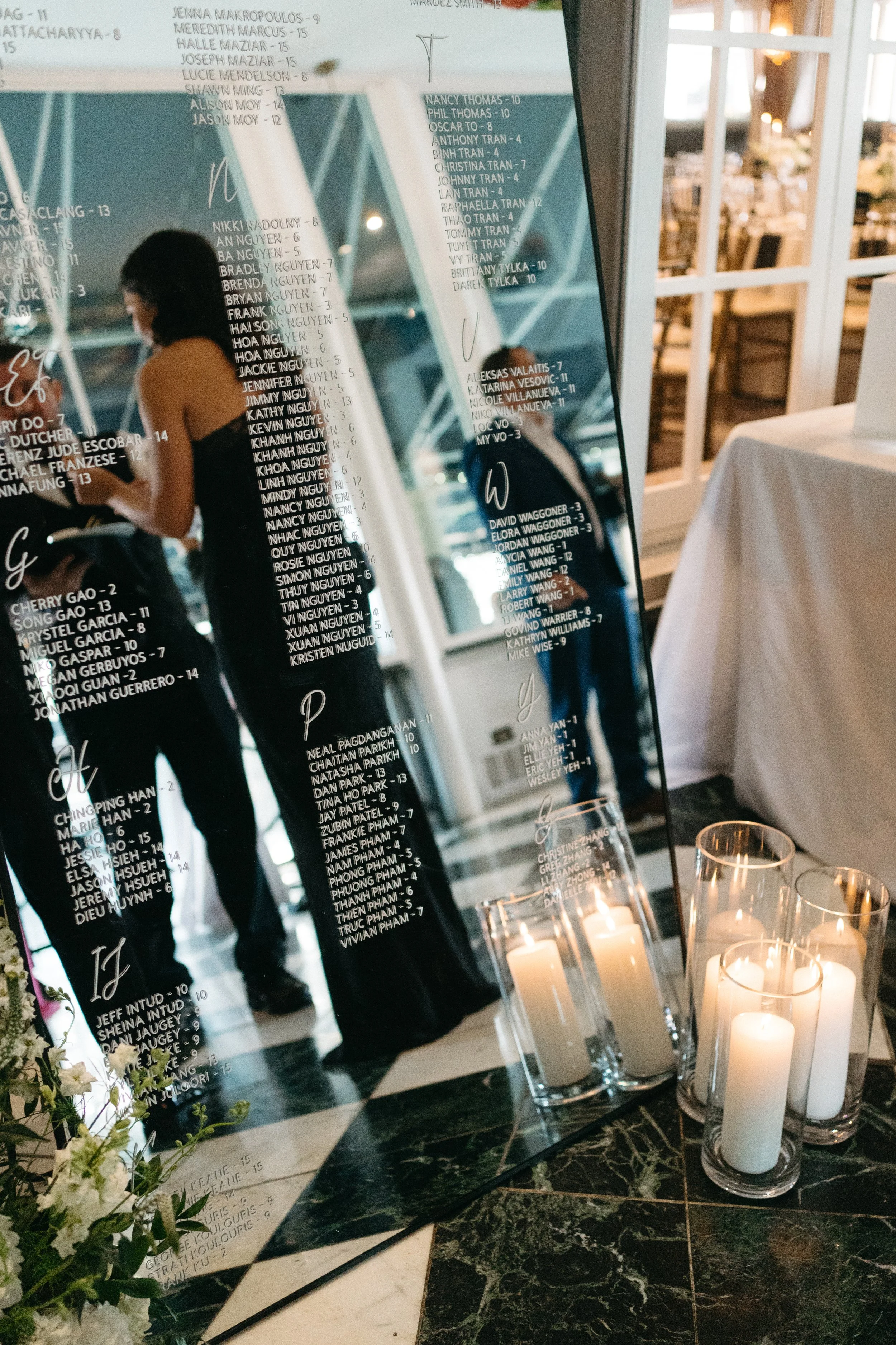 A reflective black memorial display with white printed names and candleholders with white candles, located inside a venue with white-framed glass windows and a table with a white tablecloth.