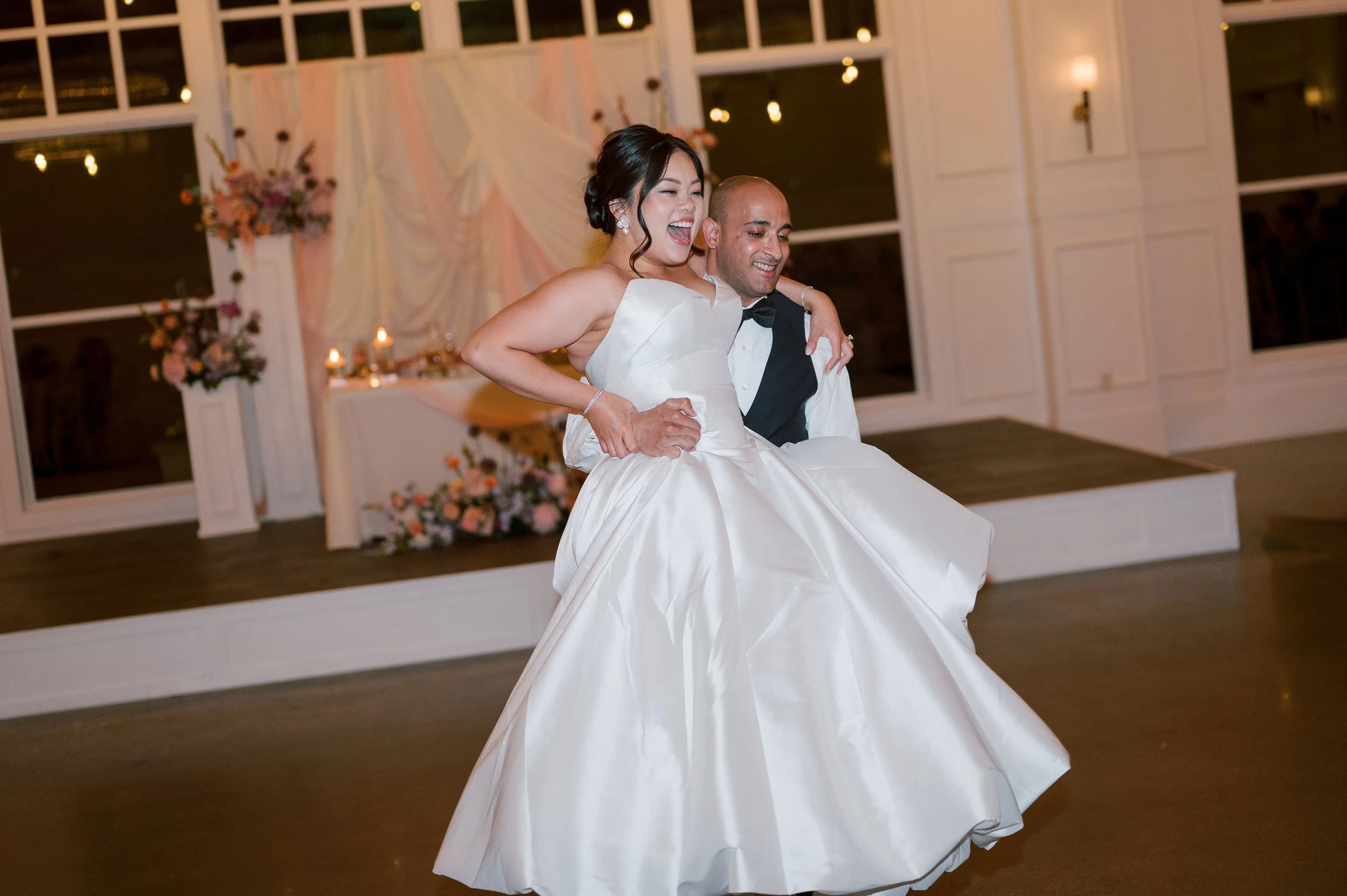A bride and groom dancing at a wedding reception. The bride is wearing a strapless wedding gown and the groom is in a tuxedo. They are smiling and appear joyful. The background features decorated windows, flowers, and candles.