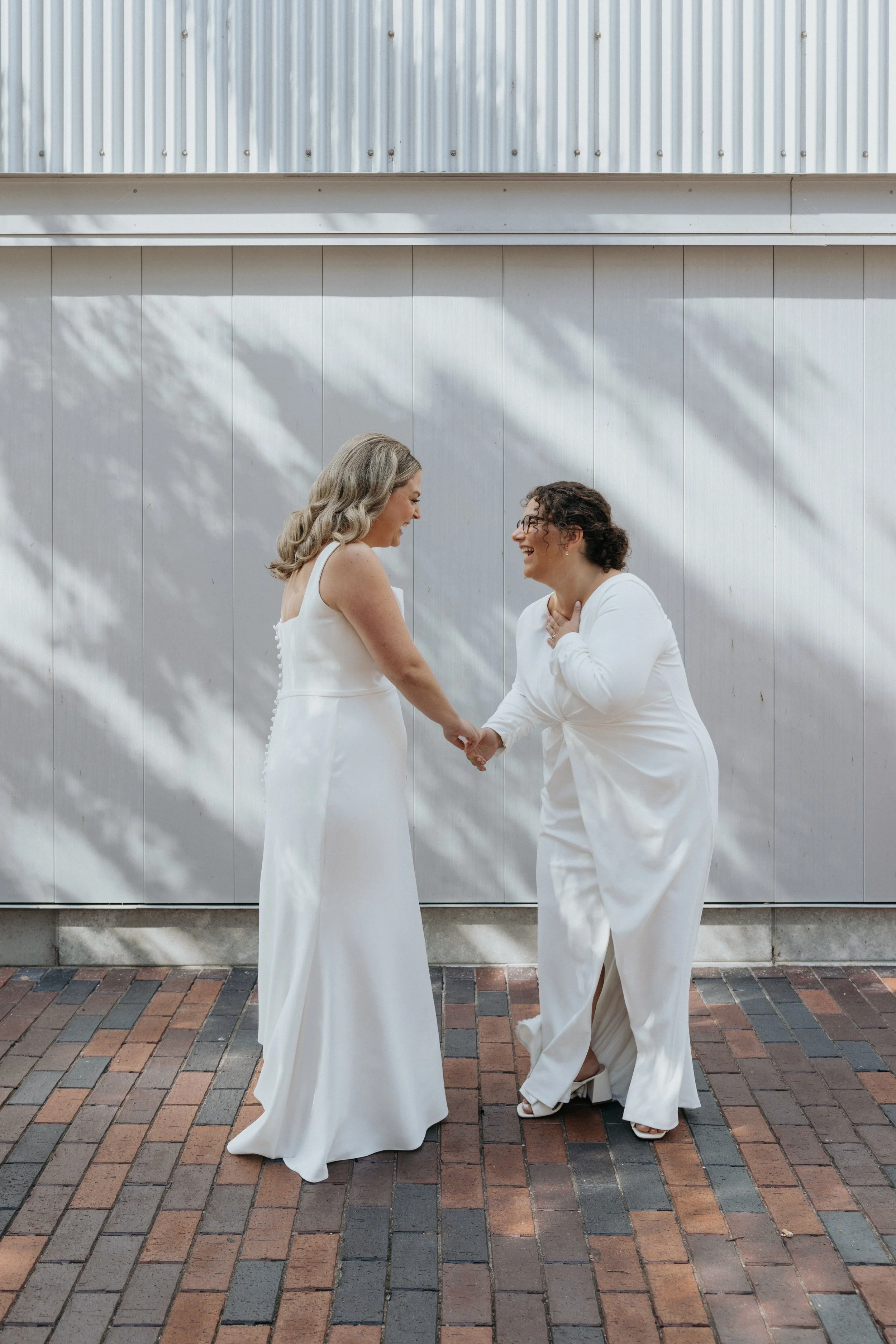 Two women dressed in white, holding hands and smiling at each other in front of a white wall with shadow patterns, on a brick sidewalk.