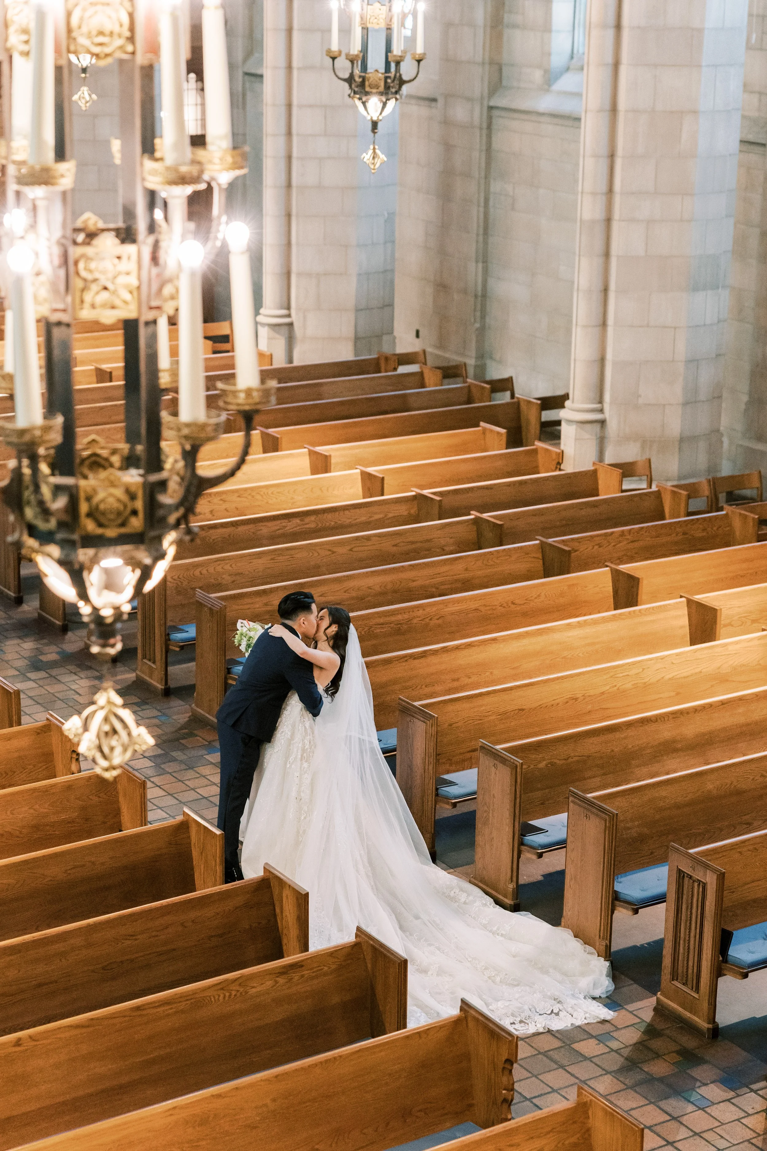 A bride and groom sharing a kiss in an empty church with wooden pews, stone walls, and chandeliers hanging from the ceiling.