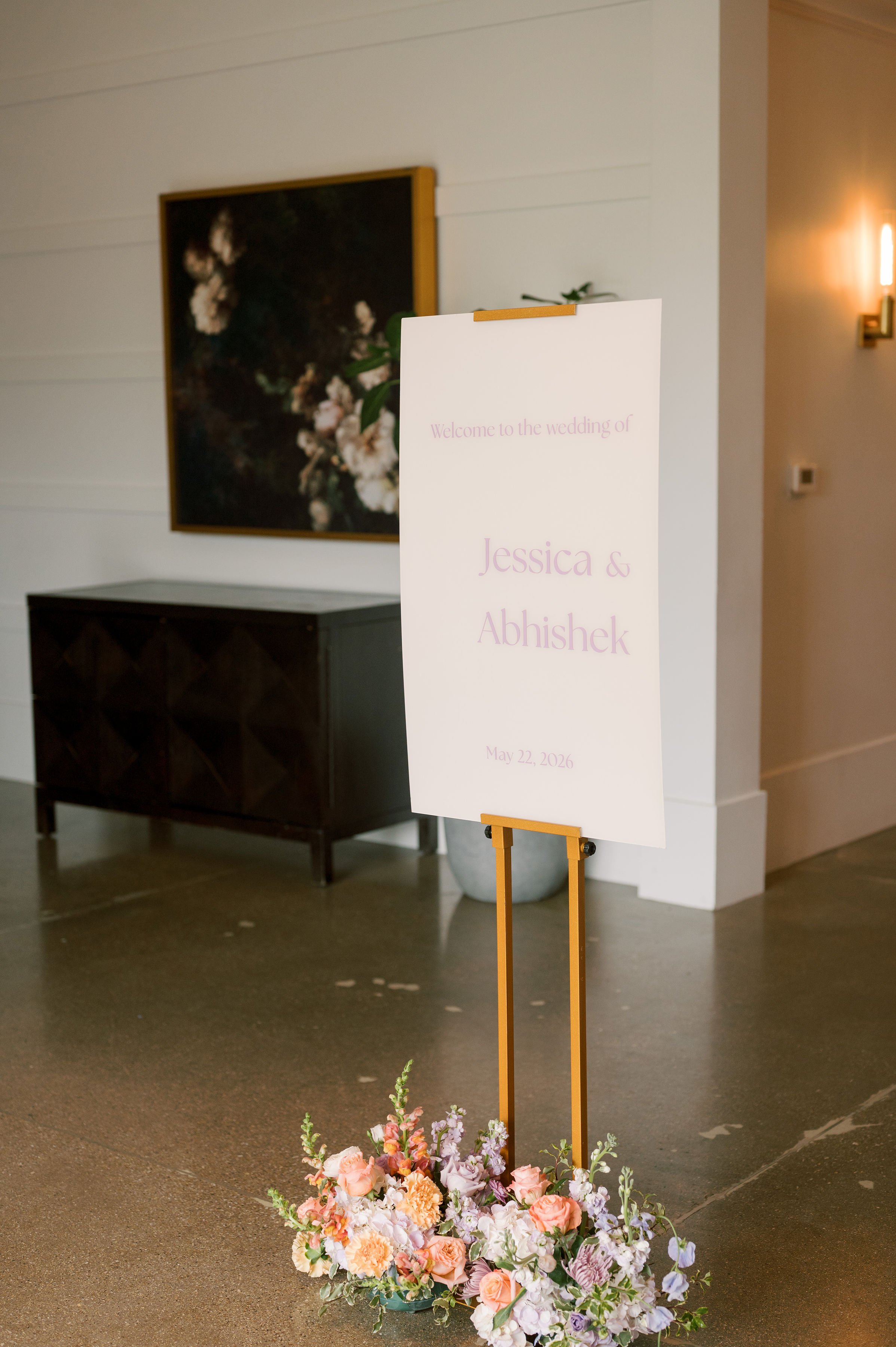 A wedding welcome sign on an easel with pink lettering, stating the names Jessica and Abishek, with flowers at the base, in an indoor venue with a black cabinet and a floral painting on the wall in the background.