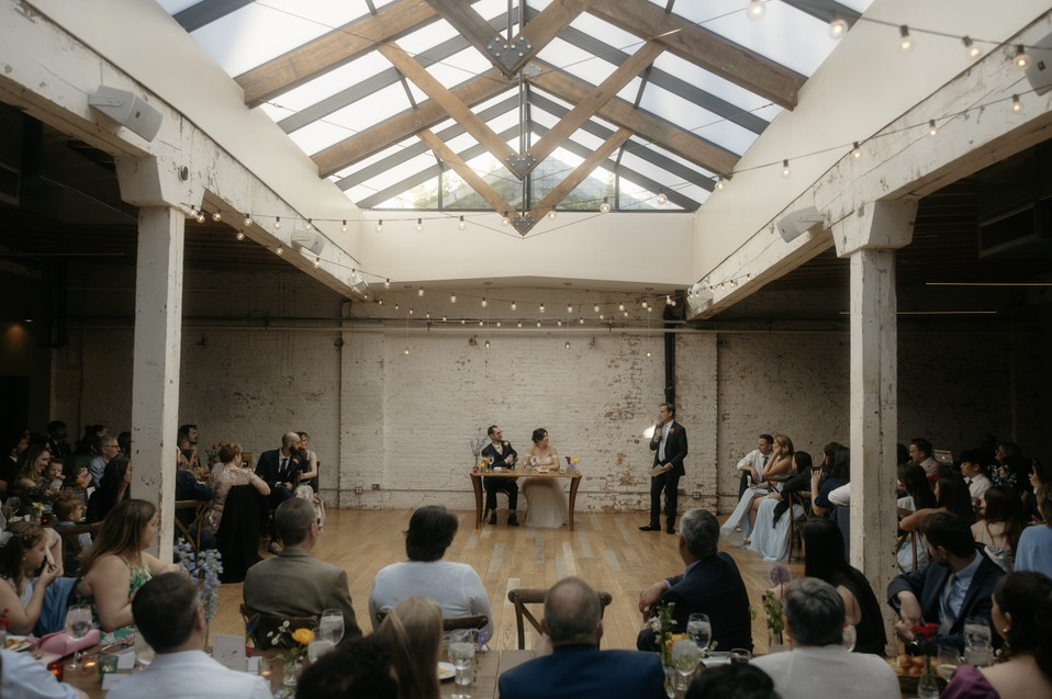 Wedding reception in an industrial-style venue with a high ceiling and string lights, featuring the bride and groom seated at a table with a wedding officiant standing nearby, while guests sit and stand around watching the ceremony.