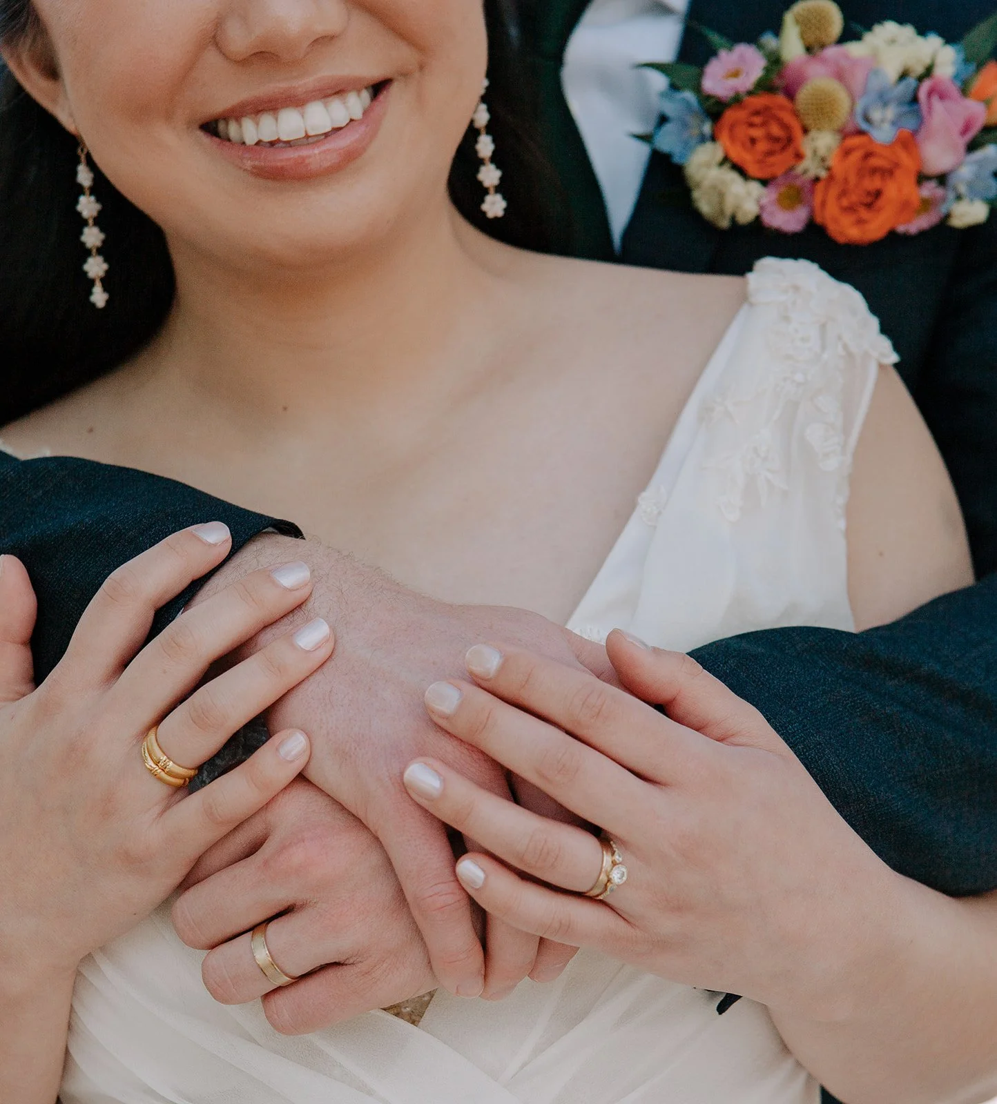 Close-up of a smiling woman in a wedding dress with multiple hands of people touching her chest and shoulder, showing rings and jewelry, and a man in a tuxedo holding a colorful bouquet of flowers in the background.