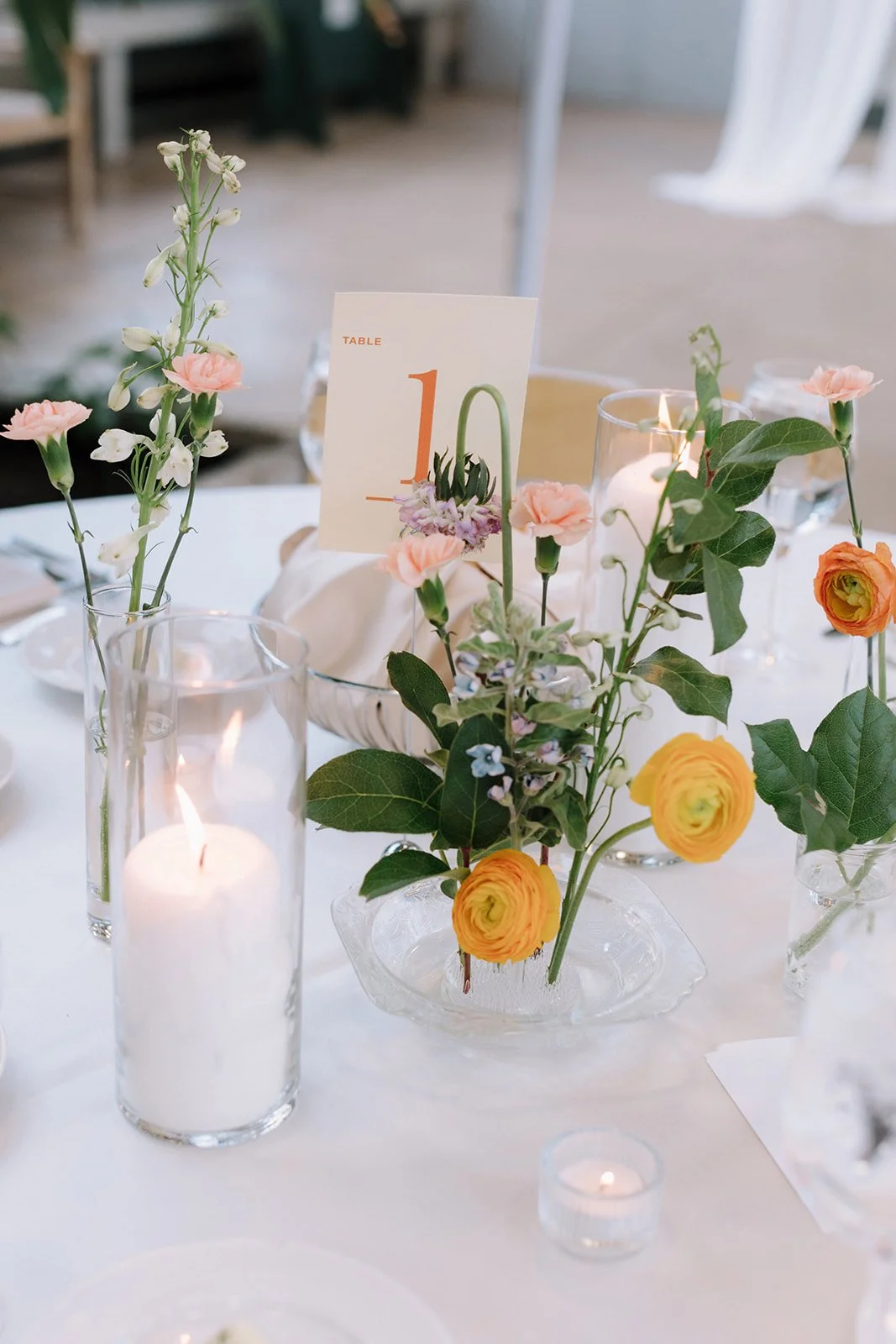 Elegant table centerpiece with pink, orange, and purple flowers in glass vases, surrounded by candles, on a white table at a wedding or special event.