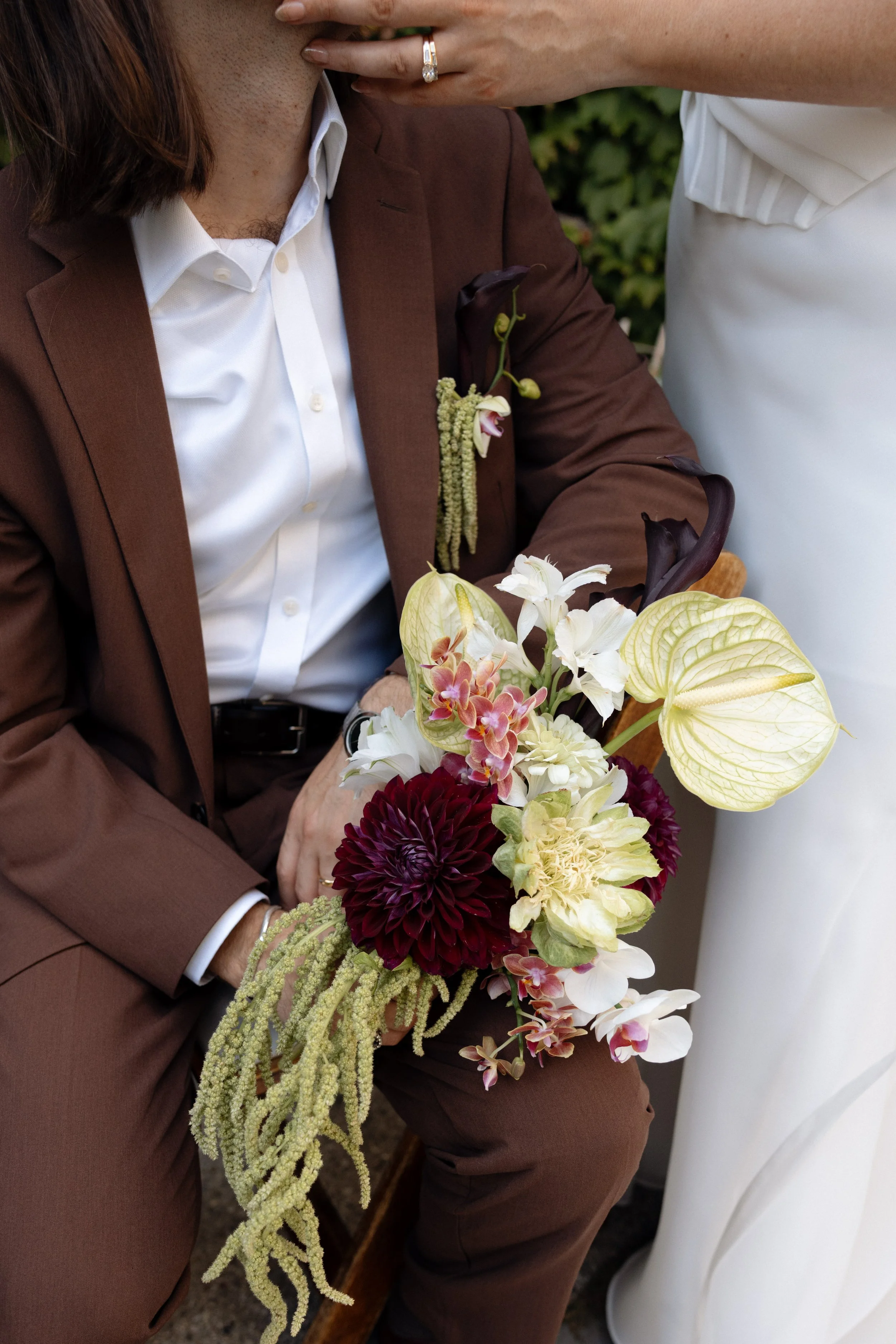 A person dressed in a brown suit holding a bouquet of flowers, including white lilies, dark maroon dahlias, and green foliage, with another person in a white dress partially visible.