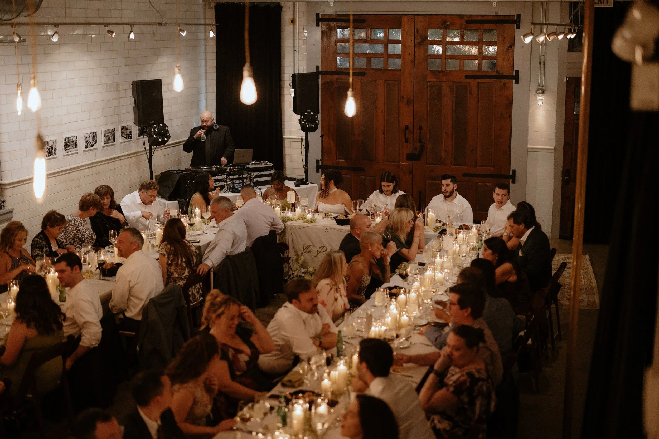 A lively indoor event with many people seated at long tables, decorated with candles, enjoying a meal. A DJ or musician with headphones is playing music near large speakers, with a rustic wooden door and a white brick wall in the background. Hanging 